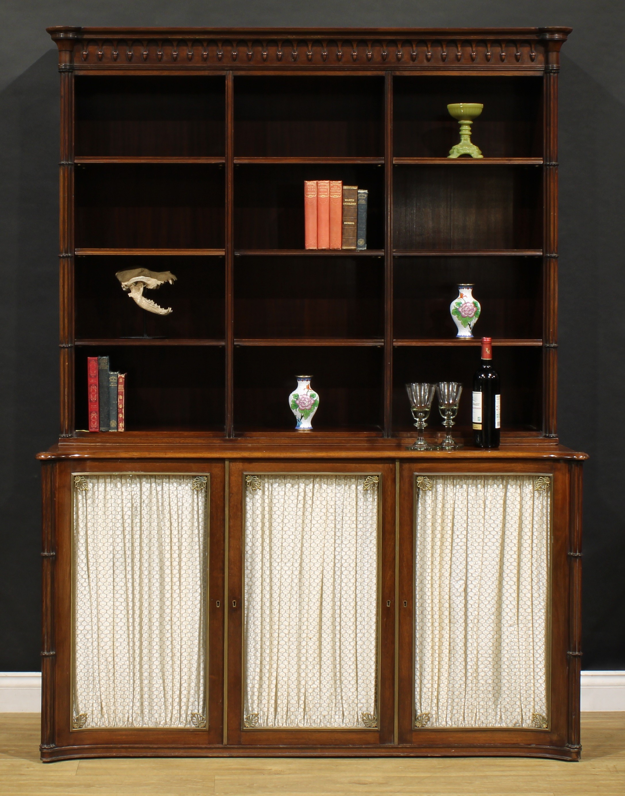 A George III Revival mahogany library bookcase, pendant frieze above three rows of adjustable shelves, the slightly incurved projecting base with three doors applied with brass grilles and lined in pleated silk, 200cm high, 147cm wide, 55cm deep, first half 20th century