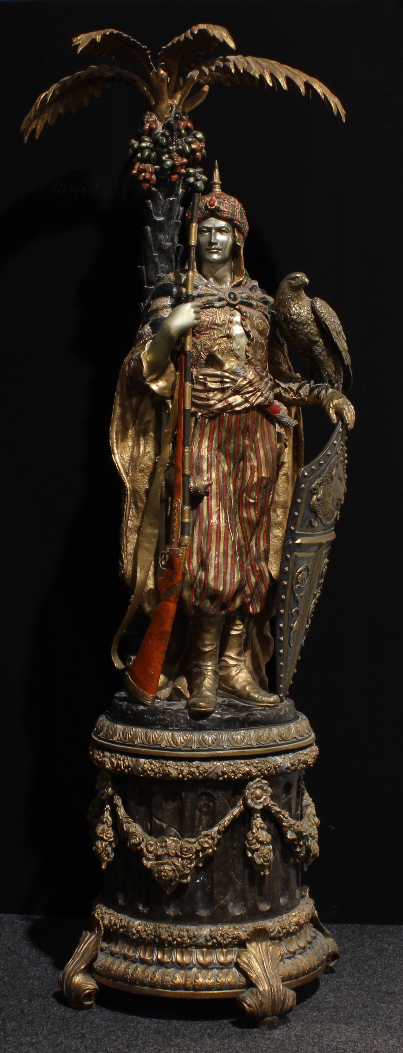 K. Namphet (contemporary) a substantial Orientalist polychrome decorated gilt and silvered bronze figure, as an Ottoman janissary, he stands with a falcon, shield and rifle, before a palm tree, Neoclassical plinth, 225cm high