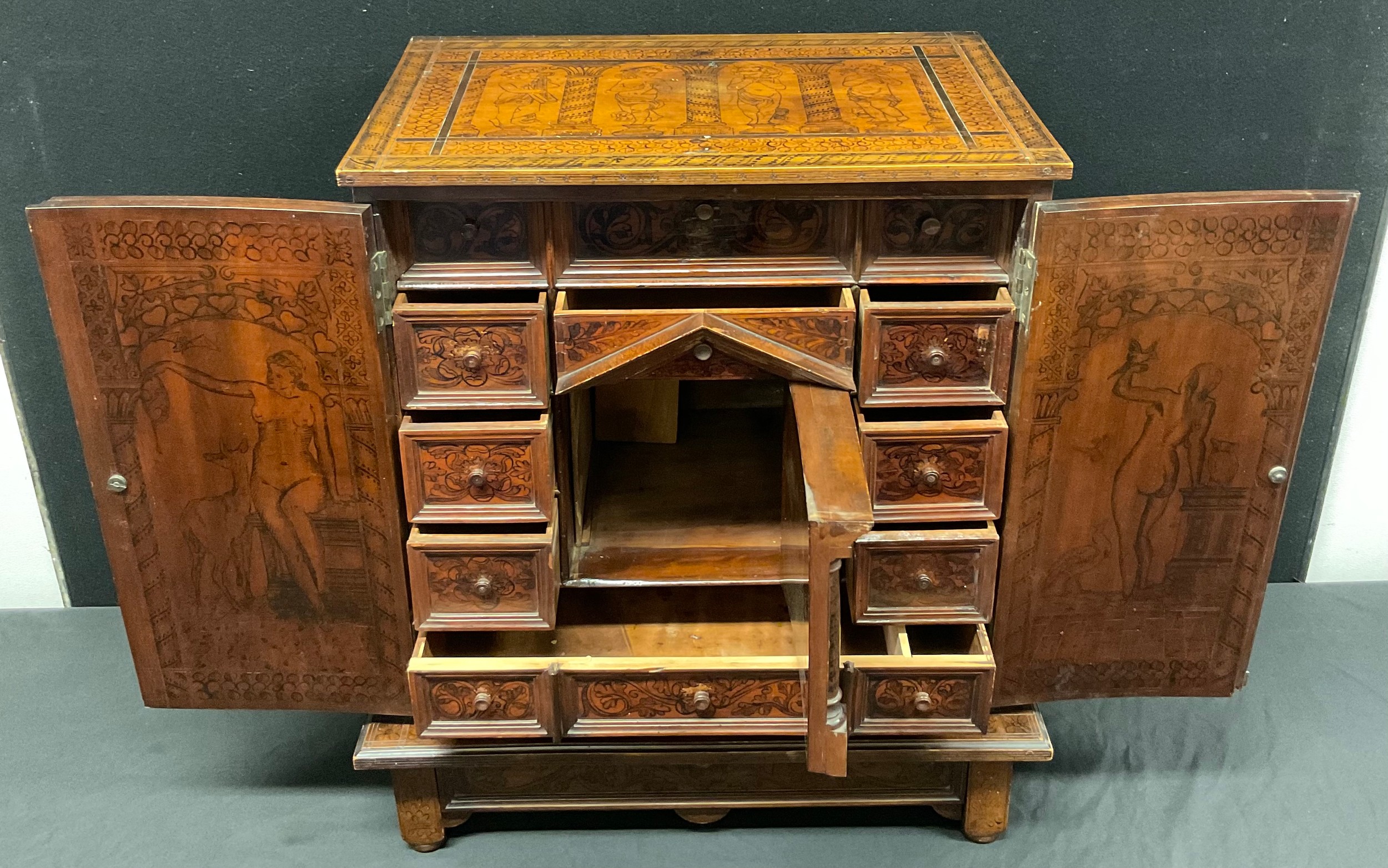 A North Italian cedar and penwork collector's cabinet on stand, hinged top above a pair of drawers enclosing an architectural interior with a cupboard centring an arrangement of wunderkammer drawers, profuesely decorated with Classical muses, nudes, putti musicians, Leda and the Swan and Diana the Huntress, the sides with silhouette figures, the base with a further long drawer, 57.5cm high overall, 44.5cm wide, 19th century
