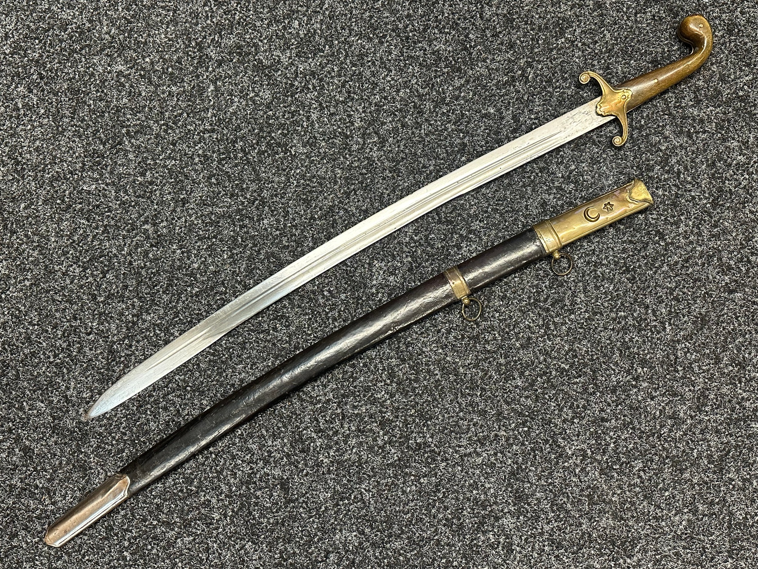 Ottoman Turkish Officers Kilij style Sword with curved double fullered blade 710mm in length. Brass crossguard. Wooden grips. Overall length 860mm. Leather scabbard with brass fittings, twin suspension rings, Star and Crecent to scabbard throat. Additional hanging ring to reverse of scabbard.