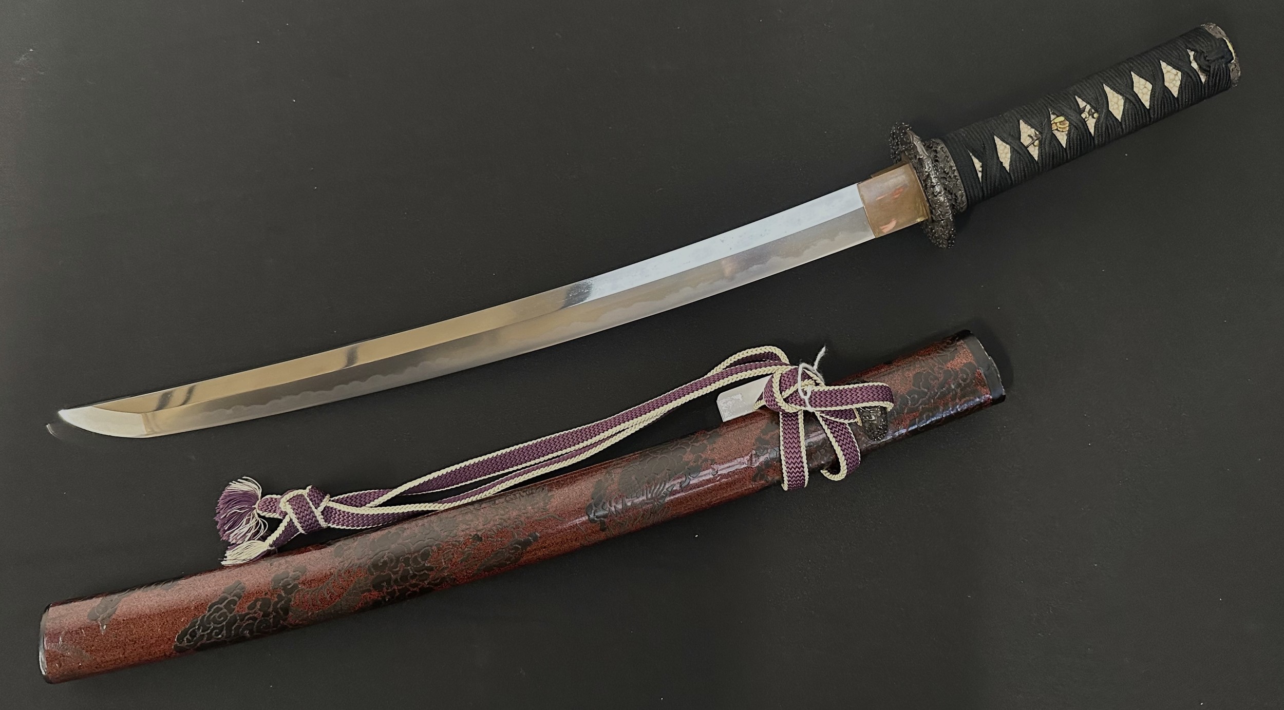 Japanese Wakizashi Sword with single edged blade 445mm in length. Good Hamon line. Signed tang. Overall length 610mm. Complete with red/black lacquered wooden saya which contains a signed Kozuka knife which is additionally maked on the blade. Has a previous Mallams Auction lot label from 2017.