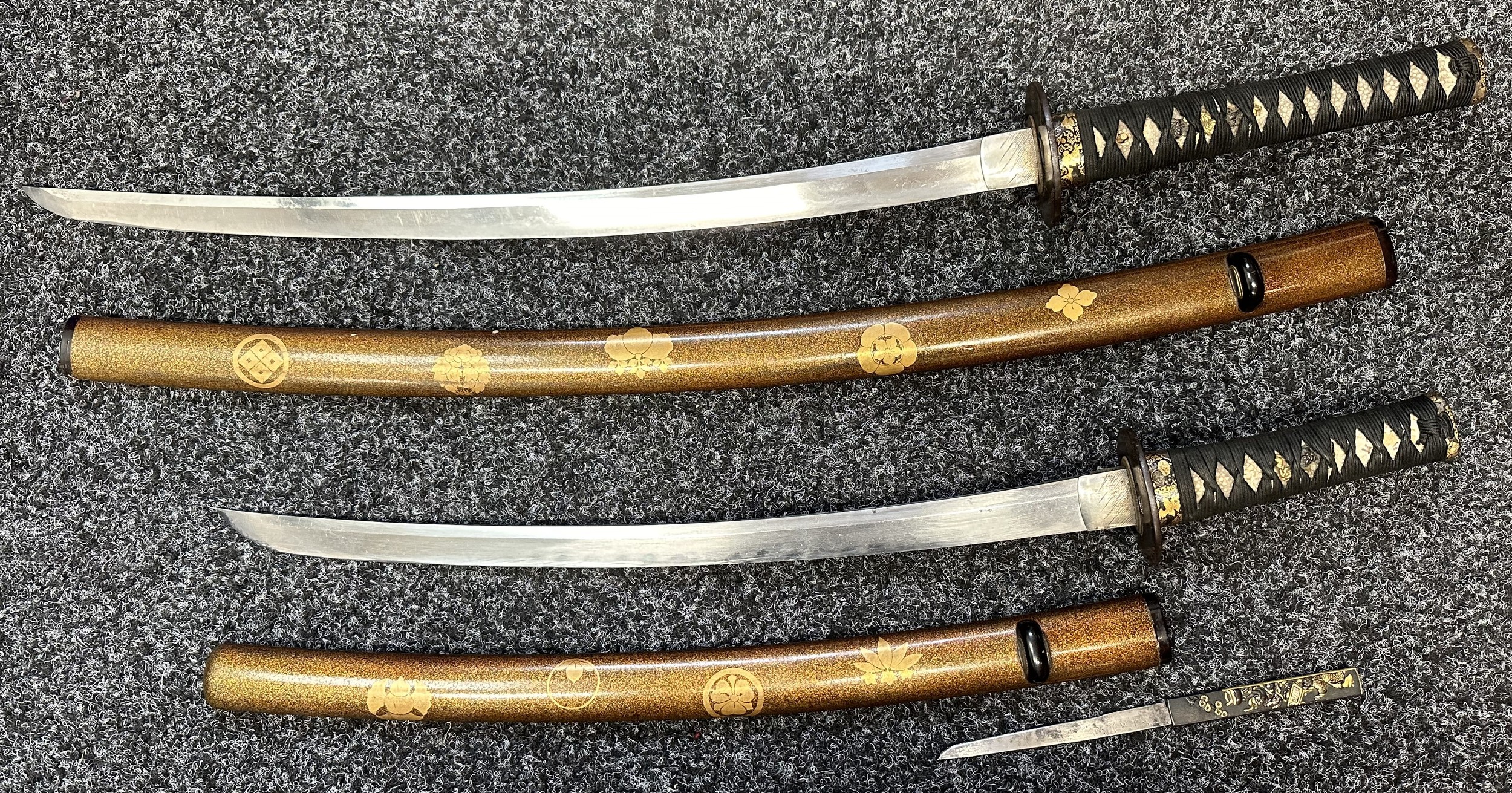 Japanese Pair Daisho Swords with complete with display stand and cloth protective bags. Larger Katana sword with single edged blade 540mm in length with Hamon line to edge, tang is not signed, ray skin grip with Menuki present, overall length 760mm. Complete with Laquered wooden Saya with gilt Mon decoration: smaller Wakizashi sword with single edged 470mm long blade with Hamon line, unsigned tang, Tsuba matches the other sword, ray skin grip with menuki, overall length 624mm. Complete with laquered wooden Saya complete with knife with signed single edged blade 120mm in length, overall length 216mm.  (2)