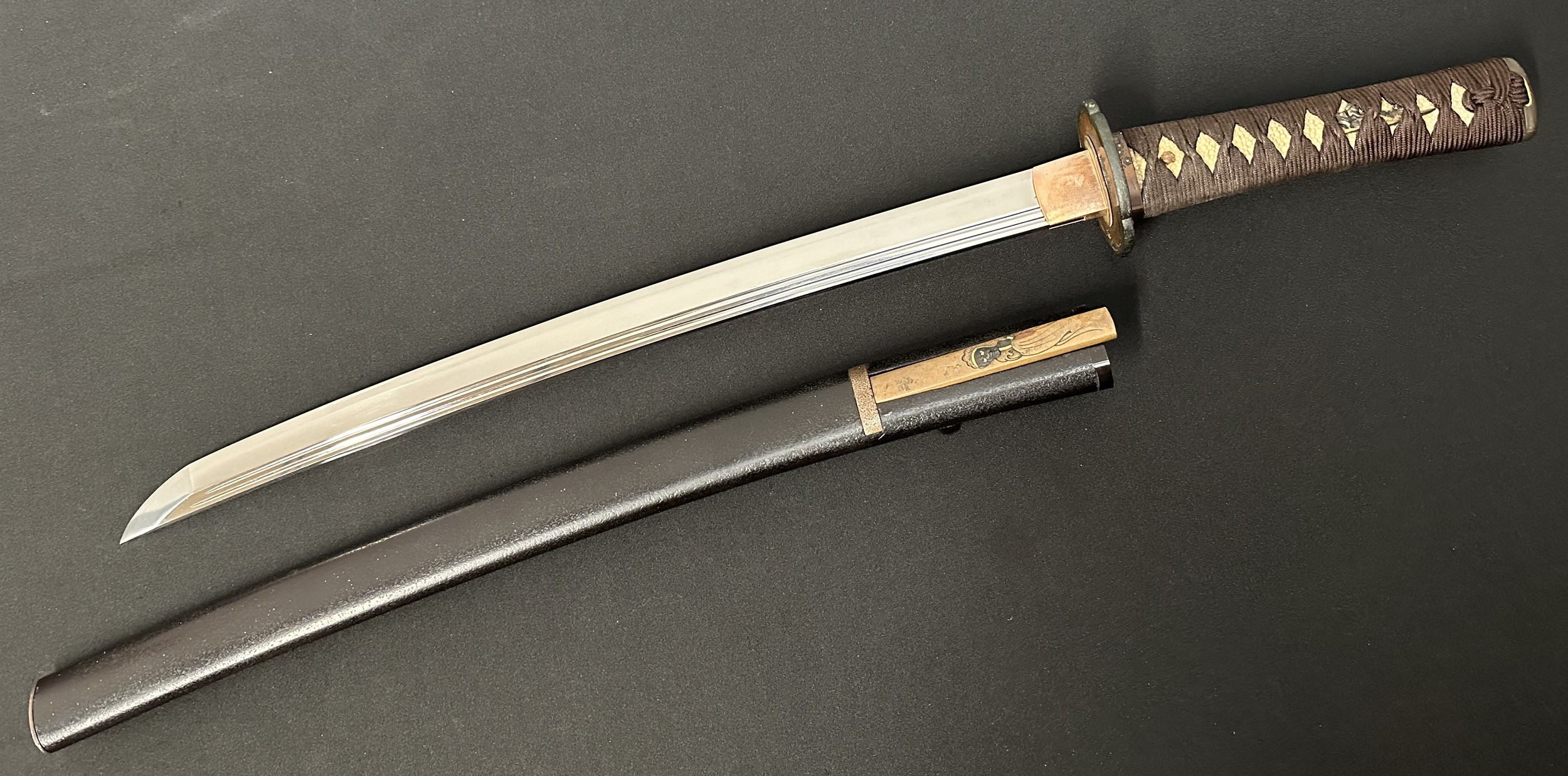 Japanese Wakizashi Sword with single edged double fullered blade 440mm in length. Hamon line to edge of blade. Two holes to tang. Overall length 610mm. Saya is complete with knife with single edged decorated blade 113mm in length with signed handle. Complete with Certificate