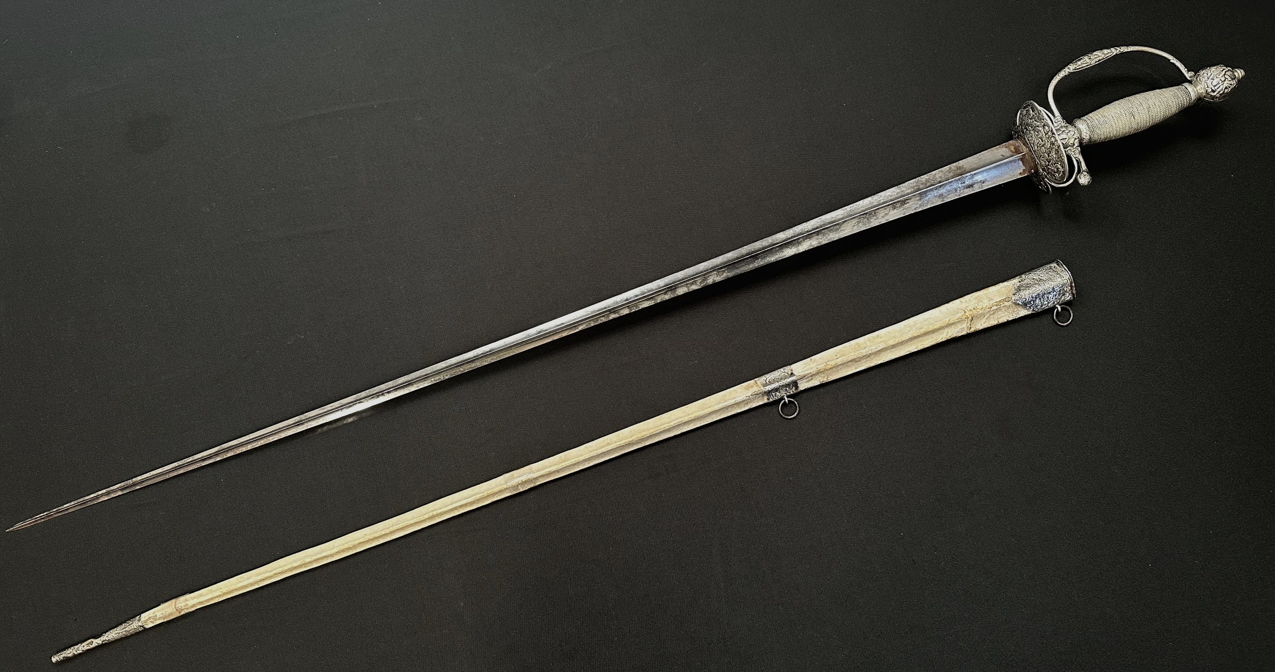 Small Sword with incurved triangular section blade 780mm in length. No makers marks. Guards featuring Classical scenes. Silver marks to D guard. Wire bound grip. Ball pommel. Overall length 950mm. Complete with white leather scabbard a/f with silver mounts.