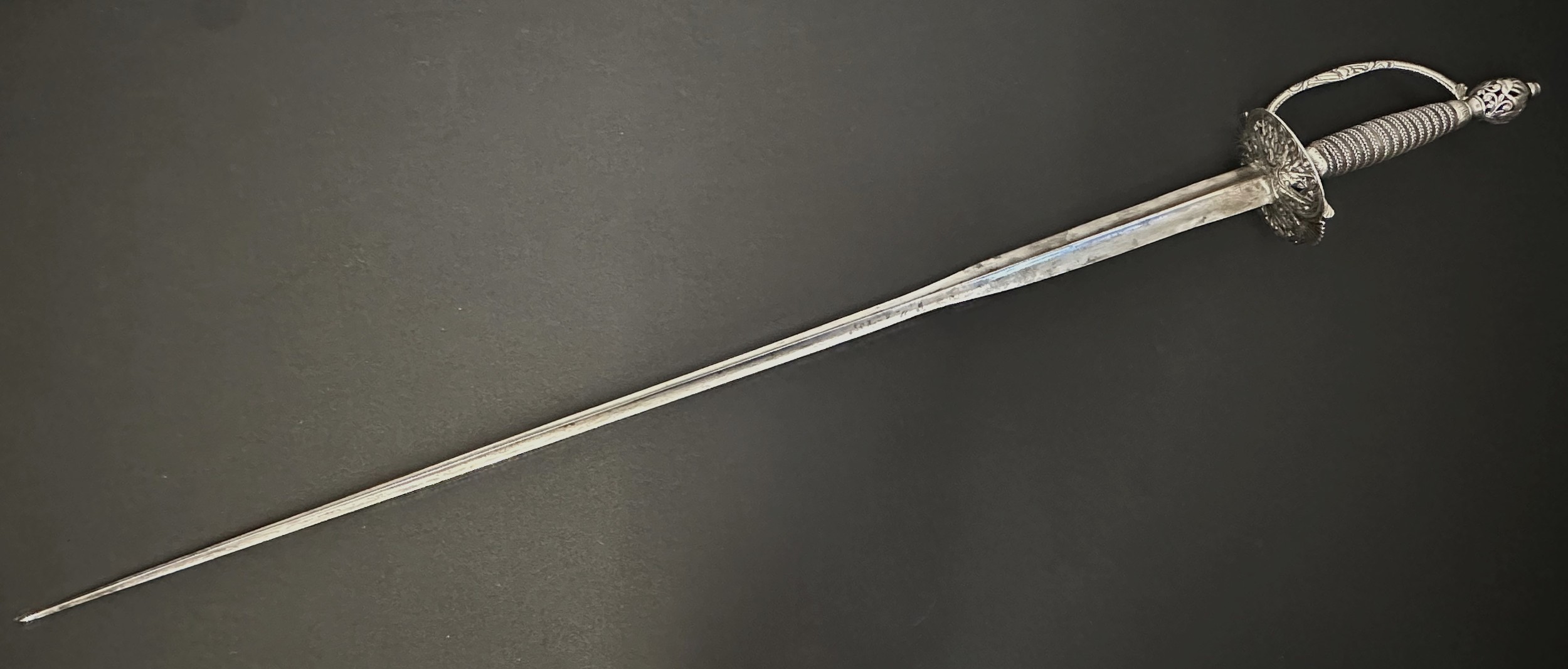 Small Sword with incurved triangular section blade 810mm in length. No makers marks. Silver peirced cirular guard with piled arms decoration and clamshells. Cross guard and D shaped guard decorated again with arms motifs. Wire bound grip. Peirced round pommel with piled arms decoration. Overall length 980mm. No scabbard.