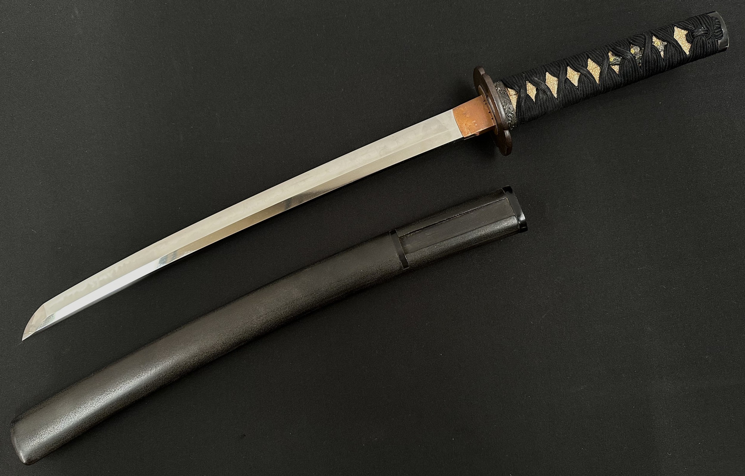 Japanese Wakizashi Sword with single edged blade 407mm in length with good Hamon line to the cutting edge. Tang is unsigned. Copper habaki & Iron Mokko Tsuba which is unsigned. Samegawa Shagreen Ray Skin grip with black Tsuka-ito cloth binding with Menuki. Overall length 585mm. Complete with brown laquered finish Saya scabbard.