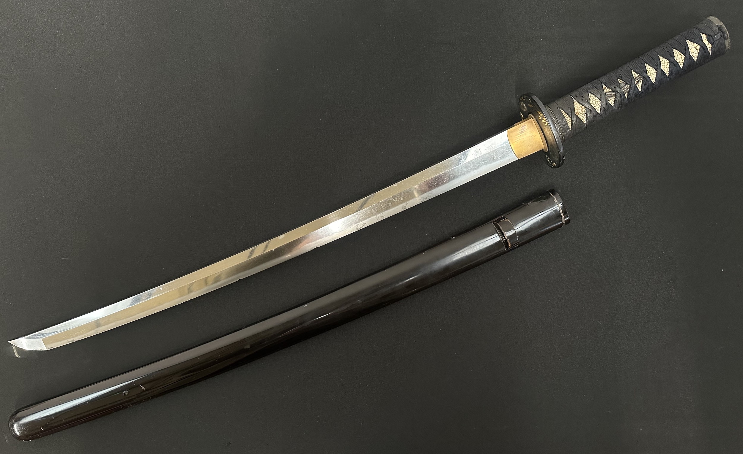 Japanese Wakizashi Sword with single edged blade 540mm in length. Hamon line to cutting edge. Some chips to edge of blade. Tang is signed. Copper plated habaki. Black Tsuba with gilt and silver decoration which is unsigned. Samegawa Shagreen Ray Skin grip with black Tsuka-ito cloth binding with Menuki present. Overall length 725mm. Complete with dark brown laquered finish Saya scabbard.