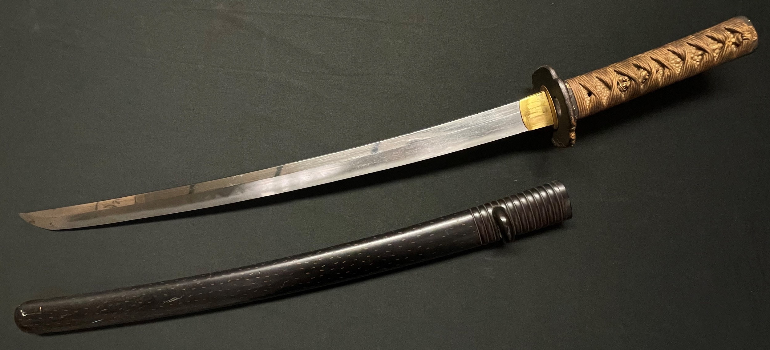 Japanese Wakizashi Sword with single edged blade 520mm in length. Hamon line to blade. Tang is unsigned but with two holes. Black iron Tsuba with gilt decoration. Fittings to the grip are again black with gilt decorations of a group of musicians. Original cloth binding over  Shagreen with Menuki in the form of Dragons. Overall length 695mm. Saya is black laquered finish with flecked gilt decoration. The release catch is decorated with gilt and black geese. War Trophy of a member of the RAF who served in the Far East, new to the market.