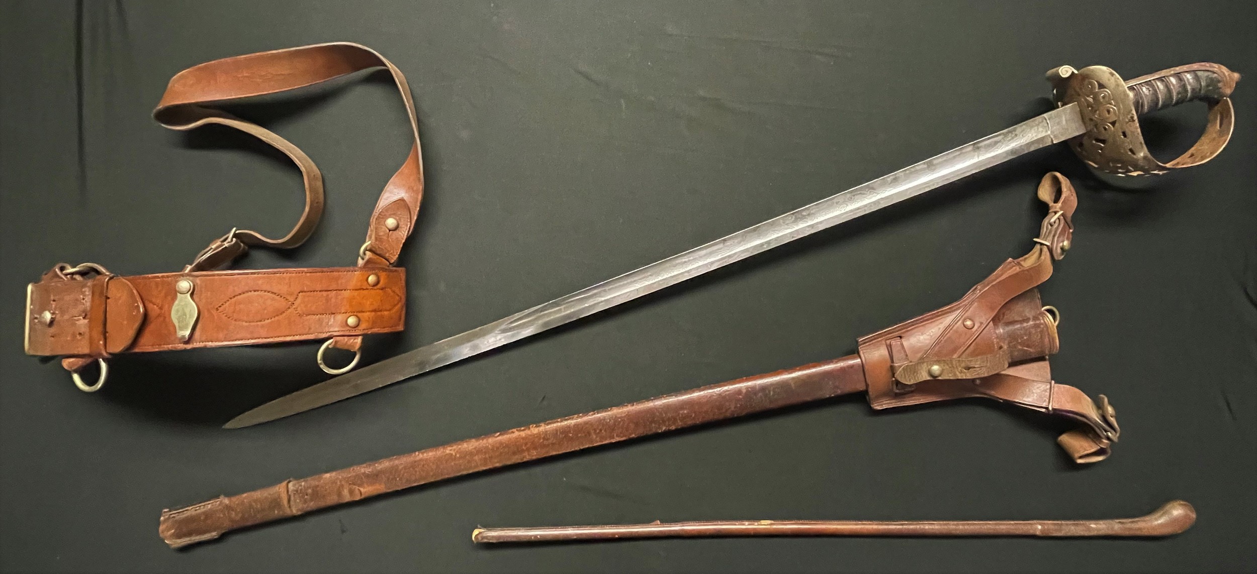 Victorian British Heavy Cavalry Officers Sword and Sam Brown Belt. Sword has decorated etched single edged fullered blade 887mm in length. Proof marked along with retailers mark "W. Anderson & Sons, 17, George Street, Edinburgh", etched panel with Queens Crown and VR, opposite side of the blade has etched panel with "Lothian & Berwickshire Yeomanry". Steel guard pierced with honeysuckle pattern design. Wire bound Shagreen grip. Overall length 104cm. Complete with leather frog and scabbard. Sam Brown belt has a name written in ink which appears to read "Rodney Thewell". In addition there is an officers brown leather covered swagger stick 71cm in length. (3)