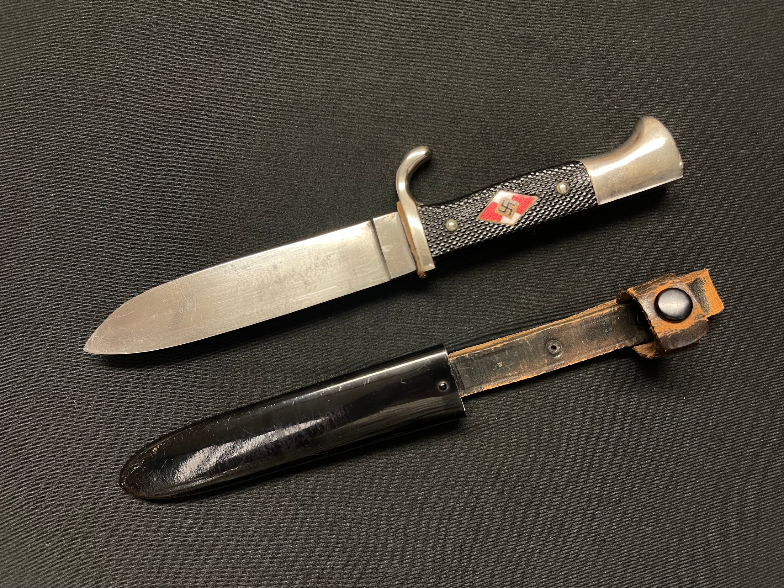 WW2 Third Reich Hitler Youth Knife with single edged blade 135mm in length, with "Blut Und Ehre!" and  transitional maker marked with "Kletterman & Moog" and "RZM M7/29 1937". Black Bakelite grip inset with enamel HJ insignia. Overall length 245mm. Leather hanger with retaining loop and press stud. Original black enamel painted finish to scabbard.