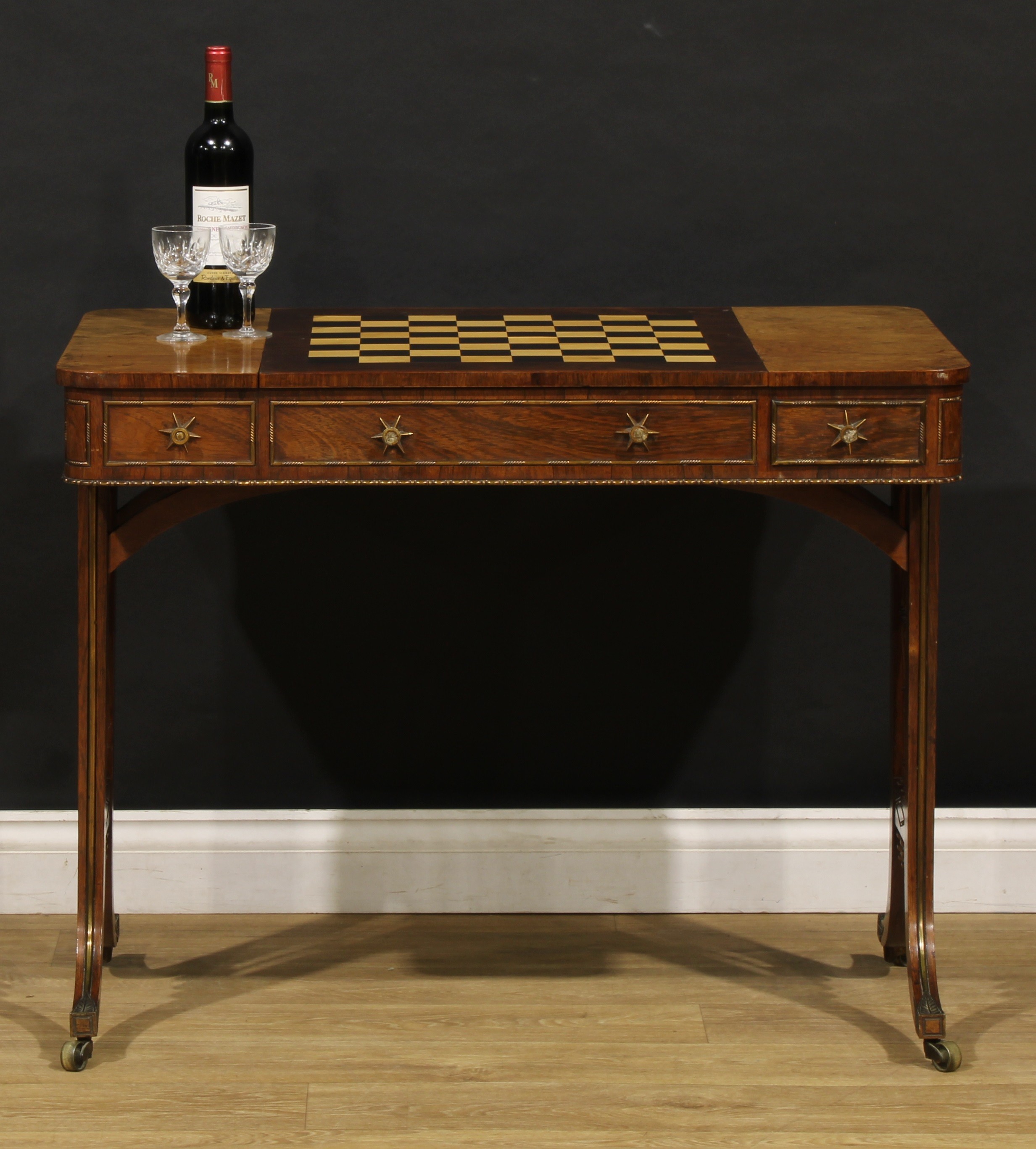 A Regency gilt metal mounted rosewood and parquetry drawing room games table, possibly Gillows of Lancaster and London, rounded rectangular top with central sliding plateau inlaid for chess and enclosing provision for backgammon, single short drawer to each long frieze, each end support with three turned spindles, sabre legs, brass casters, 73cm high, 91.5cm wide, 57.5cm deep, c.1820