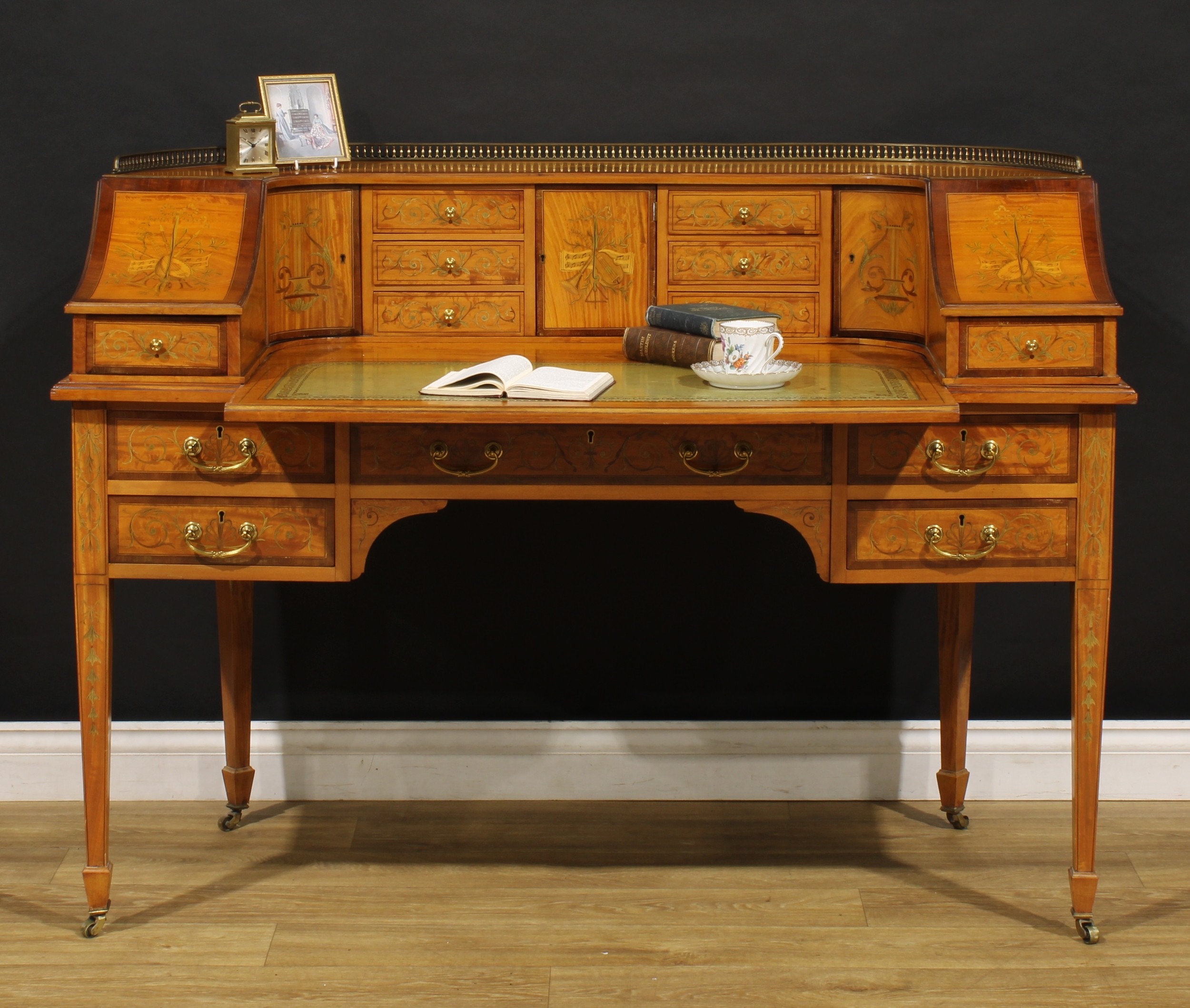 An Edwardian Sheraton Revival satinwood and marquetry Carlton House desk, shaped superstructure with brass balustrade gallery above an arrangement of drawers and doors, sliding plateau with tooled and gilt writing surface above five frieze drawers, inlaid throughout with musical trophies and scrolling foliage, tapered square legs, spade feet, brass casters, 98.5cm high, 136.5cm wide, 68cm deep, c.1905