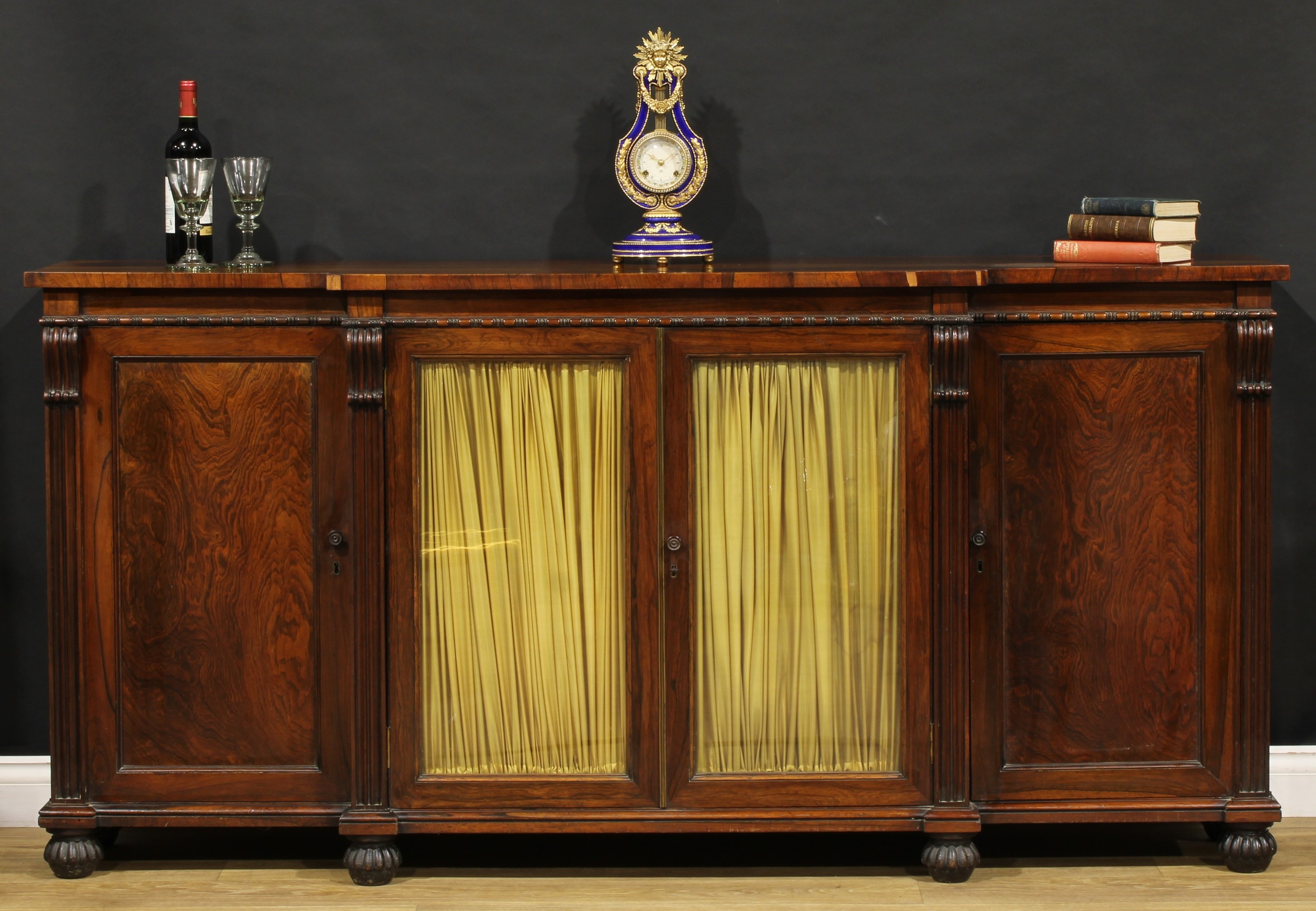 A George IV rosewood break-centre low library bookcase, oversailing top above a pair of glazed doors lined in pleated fabric, flanked by further panel doors, plinth base, 97cm high, 198cm wide, 42cm deep, c.1825