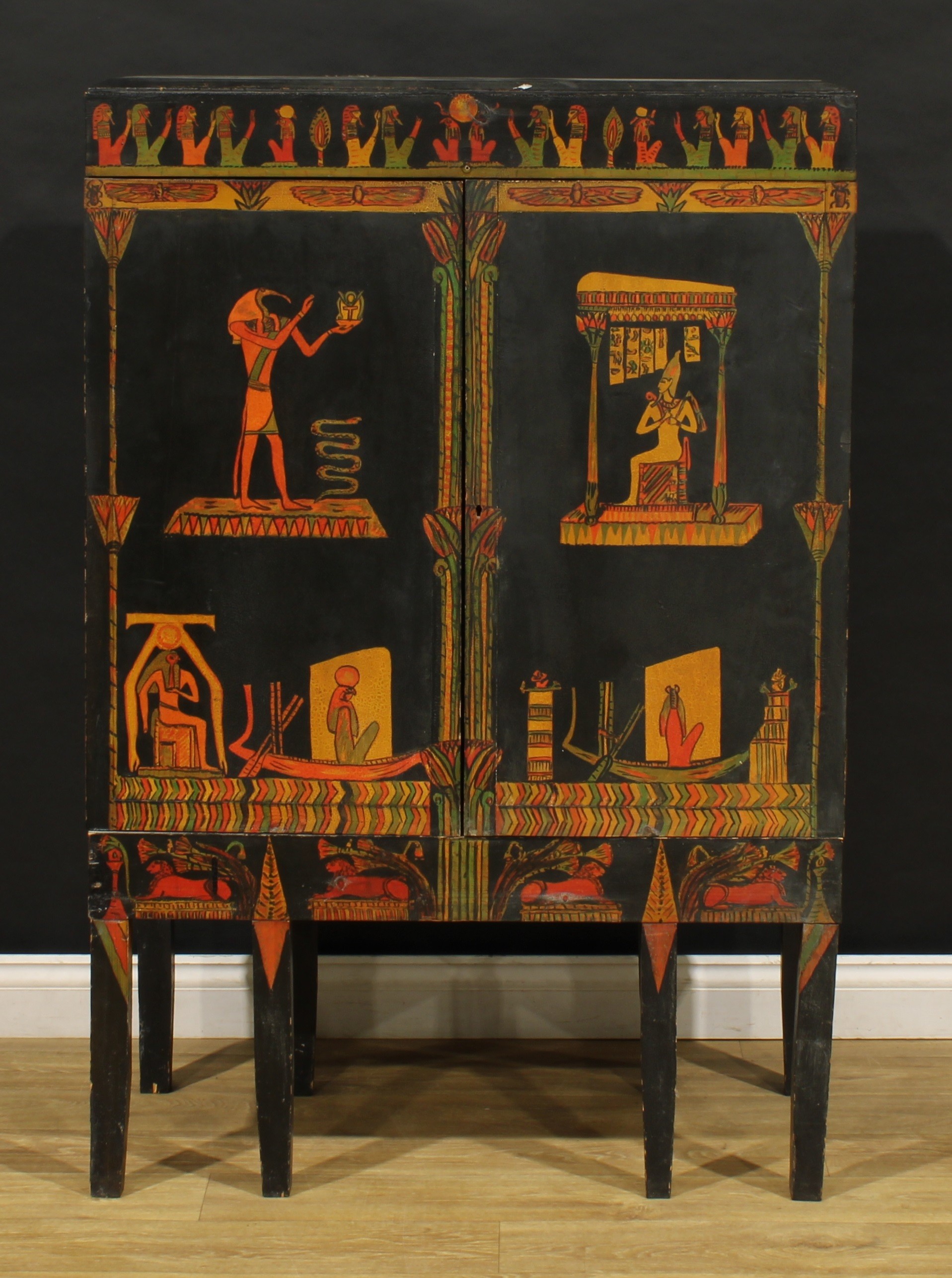 An Egyptian Revival design side cabinet, ebonised and polychrome decorated in the manner of The Great Virgil’s Disembodied Princess, the pair of panel doors enclosing pigeonholes and further small doors, 137cm high, 91cm wide, 45.5cm deep, mid-20th century