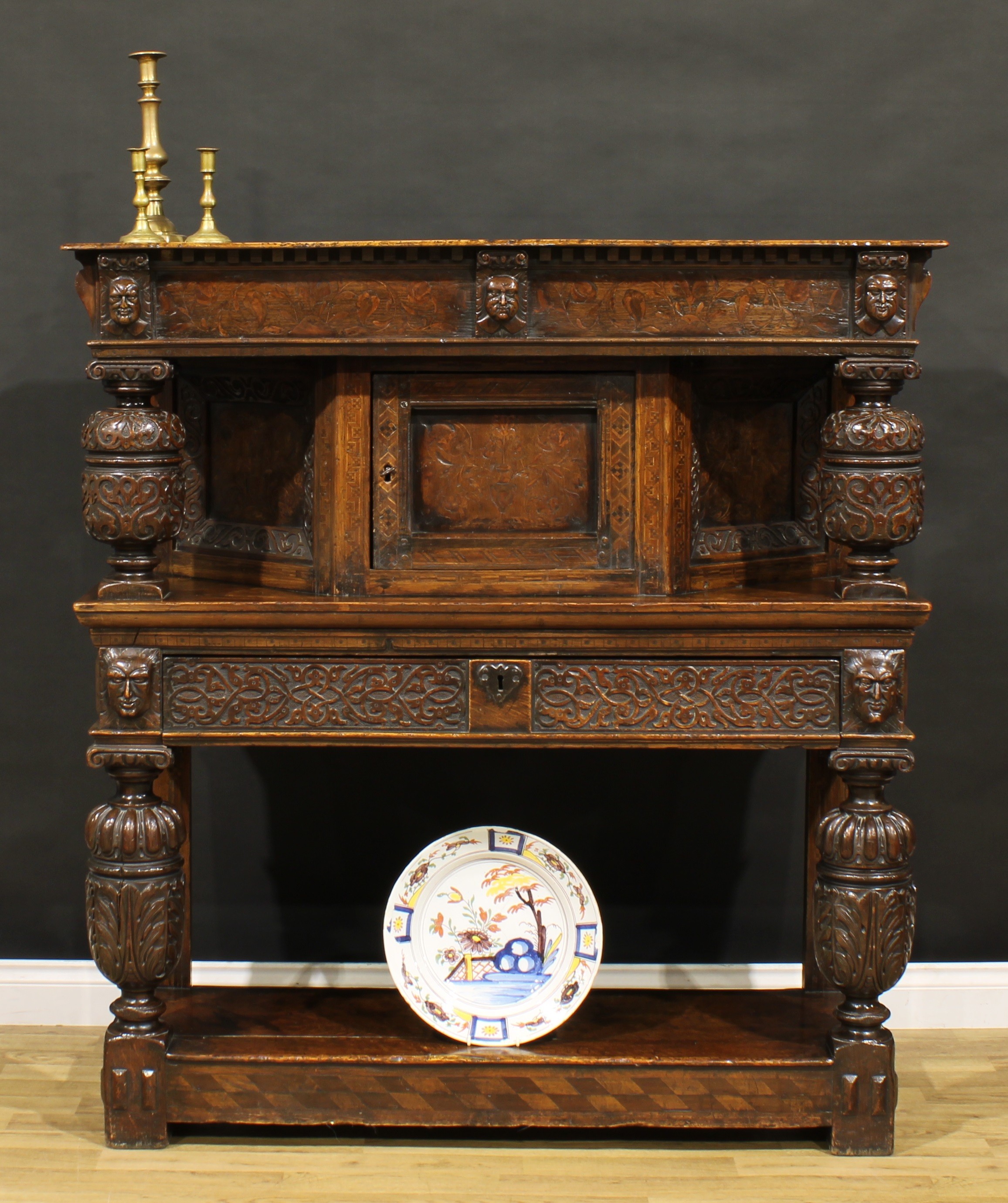 A 17th century style oak and marquetry duodarn, rectangular top with dentil capital and a frieze inlaid with meandrous flowering stems and applied with masks, above a panel door flanked by pillars, turned and carved in the Elizabethan taste, the base with a long frieze drawer carved with blind fretwork, open undertier, 144cm high, 135cm wide, 45cm deep, a later construction incorporating some period elements