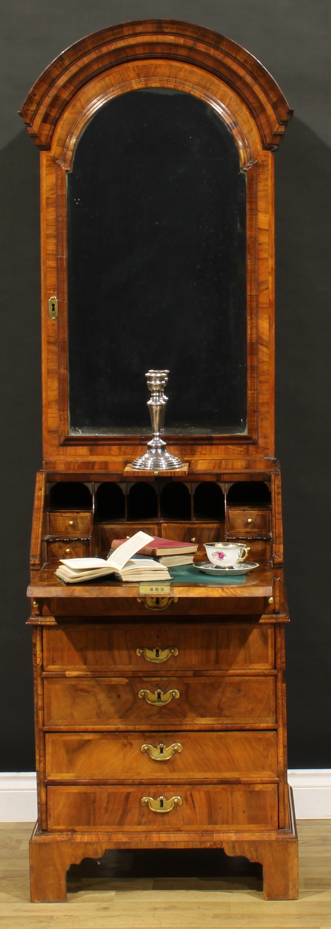 An 18th century walnut bureau book cabinet, of narrow proportions, dome top with bevelled glazed door enclosing an architectural interior with two adjustable shelves, a small door, small drawers and pigeonholes, the projecting base with fall front enclosing further small drawers and pigeonholes above five long drawers, the upper and second divided by a slide, skirted base, bracket feet, 188.5cm high, 57cm wide, 46cm deep