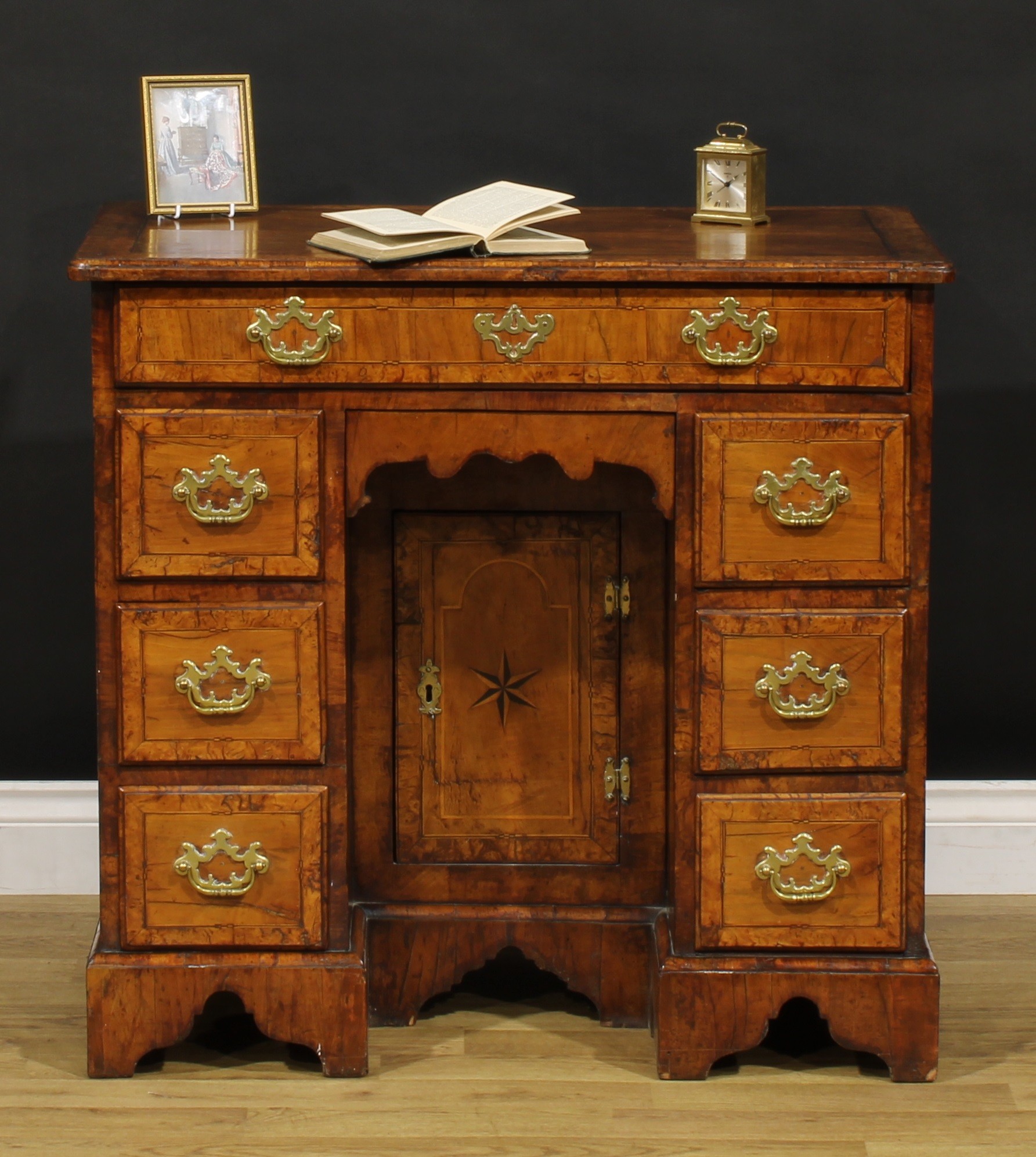 A George II walnut kneehole desk, burr-banded quarter-veneered rectangular top with moulded edge above an arrangement of drawers, brass swing handles, batwing backplates, door to niche inlaid with a six-pointed star and enclosing a shelf, bracket feet, 76.5cm high, 81cm wide, 46cm deep, c.1750