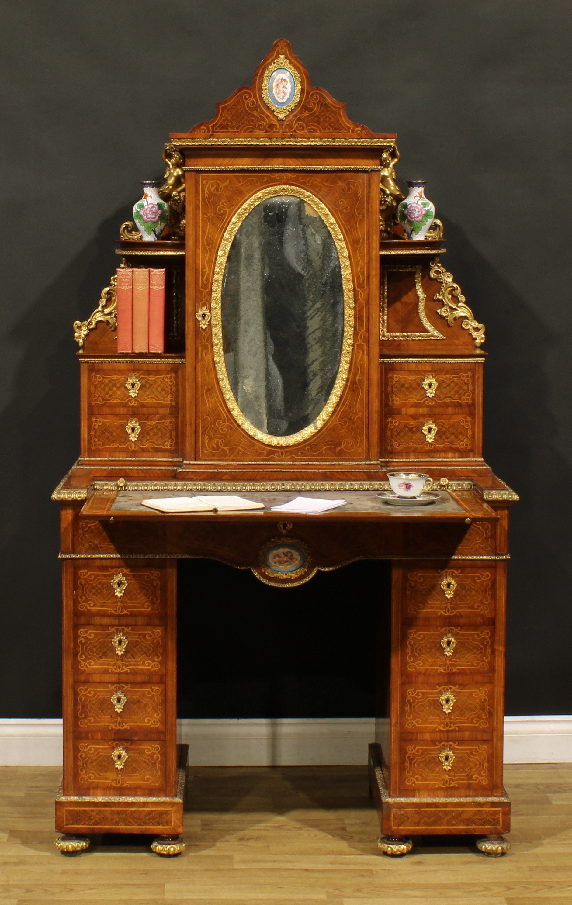 A Louis XV Revival gilt metal and porcelain mounted kingwood and marquetry exhibition-type twin pedestal escritoire desk or bonheur du jour, shaped superstructure central by a door with inset oval looking glass, enclosing two shelves, flanked by further open shelves and small drawers, the base with retractable plateau with inset tooled and gilt writing surface, above a long frieze drawer, four further drawers to each pedestal, plinth base, turned forefeet, 181cm high, 99.5cm wide, 45.5cm deep, c.1900
