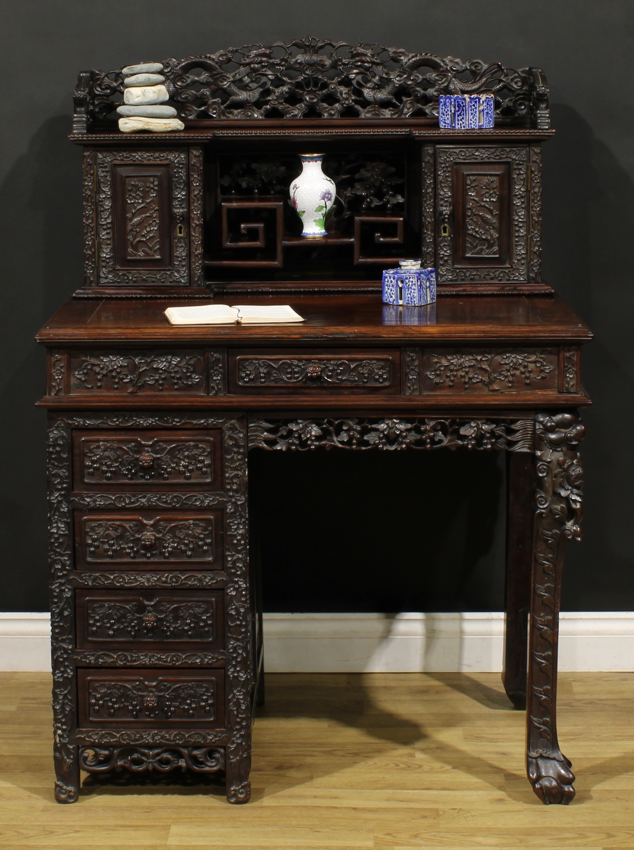 A Chinese export hardwood desk, inverted break-centre superstructure with shaped three-quarter gallery pierced and carved with dragons chasing a pearl, above a stepped three-tier plateau flanked by panel doors, carved with a bird amongst blossoming prunus, rectangular top above a central frieze drawer, the kneehole flanked by four further drawers, 127.5cm high, 91cm wide, 59.5cm deep, early 20th century