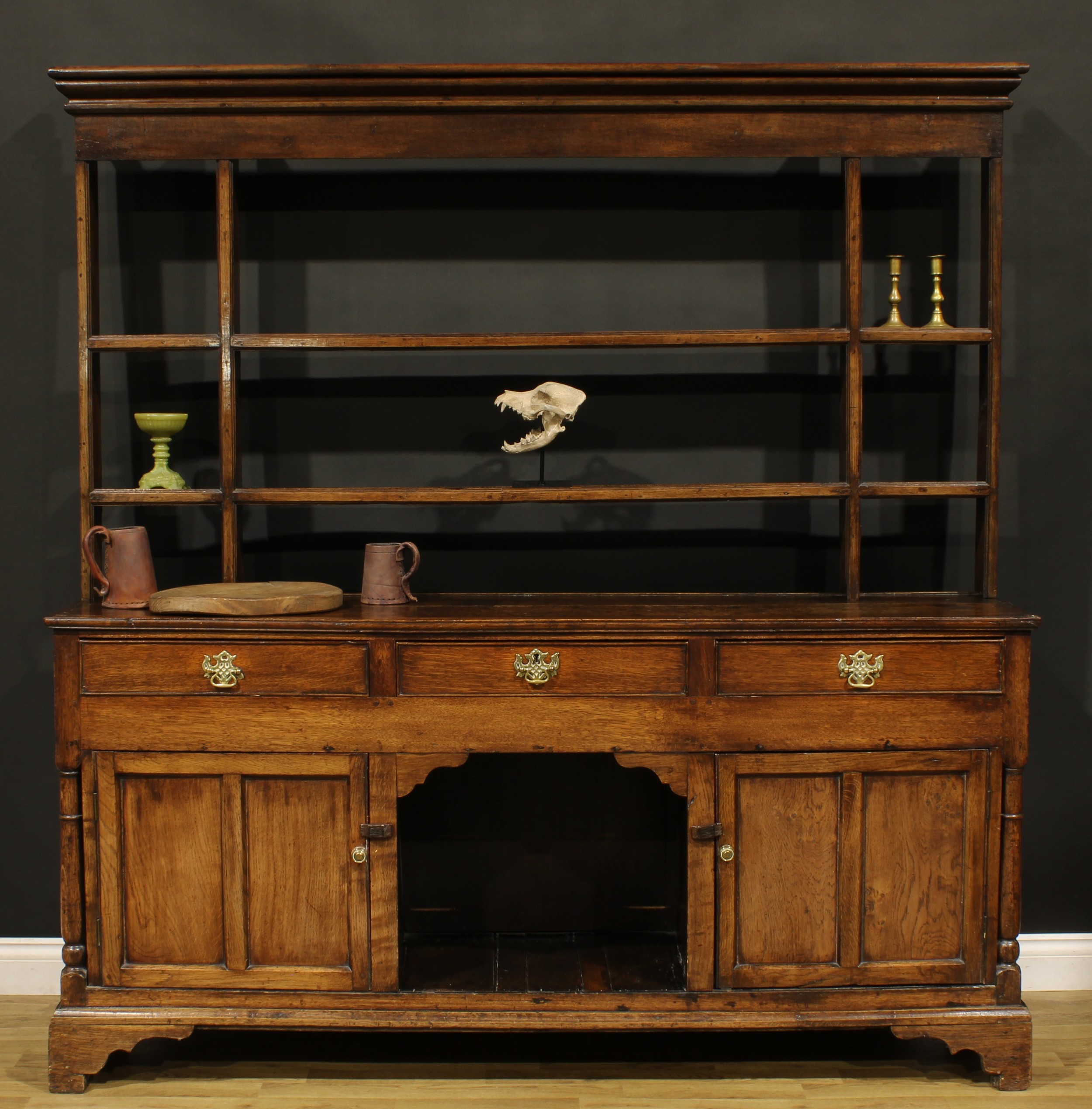 A George III oak dog kennel dresser, moulded cornice above a pair of plate racks flanked by open niches, the projecting base with three short drawers above a pair of panel doors centred by an open potboard, skirted base, bracket feet, 188cm high, 184.5cm wide, 47cm deep, c.1800