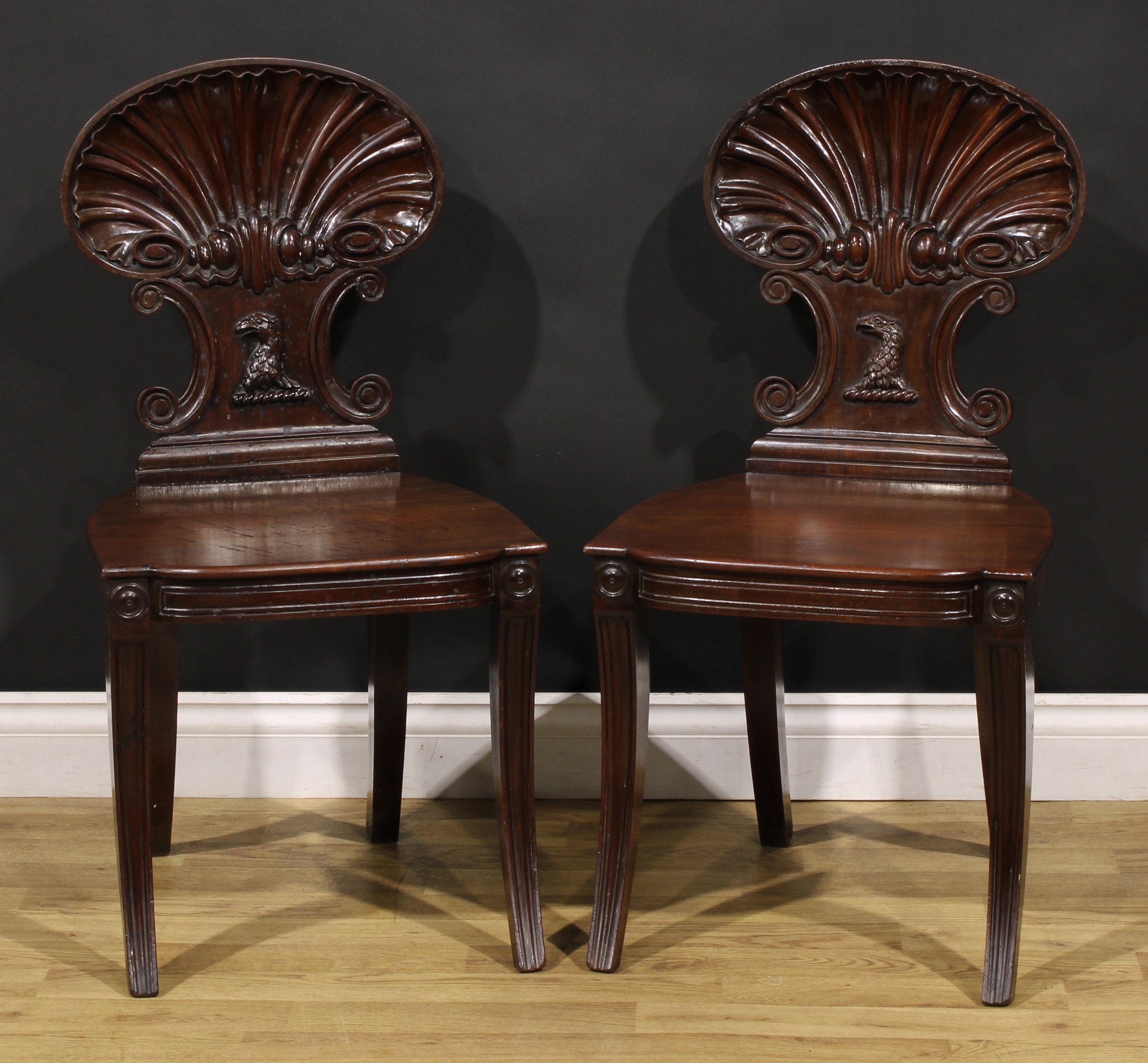 A pair of Post-Regency mahogany hall chairs, in the manner of Gillows of Lancaster and London, each with a shaped back carved with a shell, C-scrolls and applied with an eagle head, sabre legs, 85.5cm high, 43.5cm wide, the seat 39cm deep, 19th century