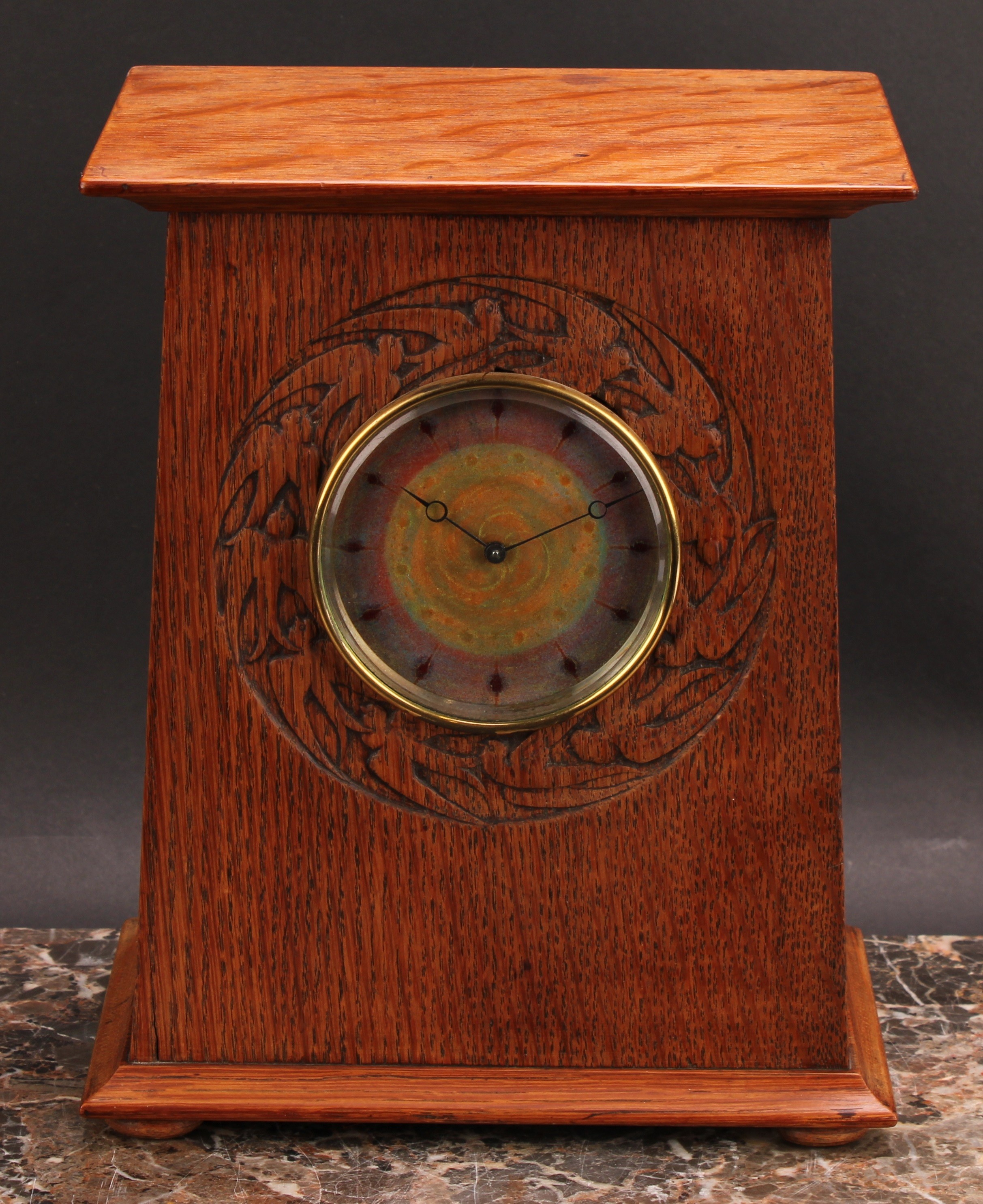 An Arts and Crafts oak mantel timepiece, 9cm circular dial decorated in mottled tones of orange and purple blush enamel, eight-day movement, the case with oversailing pediment, carved with a bezel band of swallows, bun feet, 26cm high, 21.5cm wide, c.1910