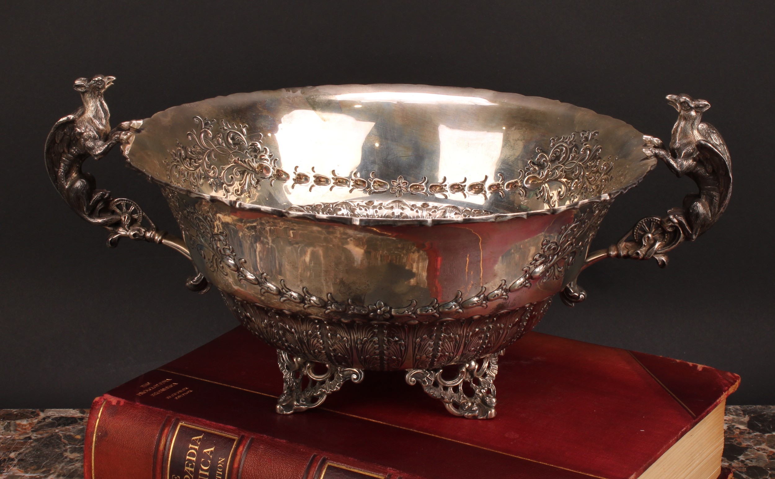 A silver two-handled table centre, of substantial gauge, chased with stiff leaves and bell husks, the handles cast as winged mythical beasts, four pierced bracket feet, 39.5cm wide over handles, London 1895, 1628g