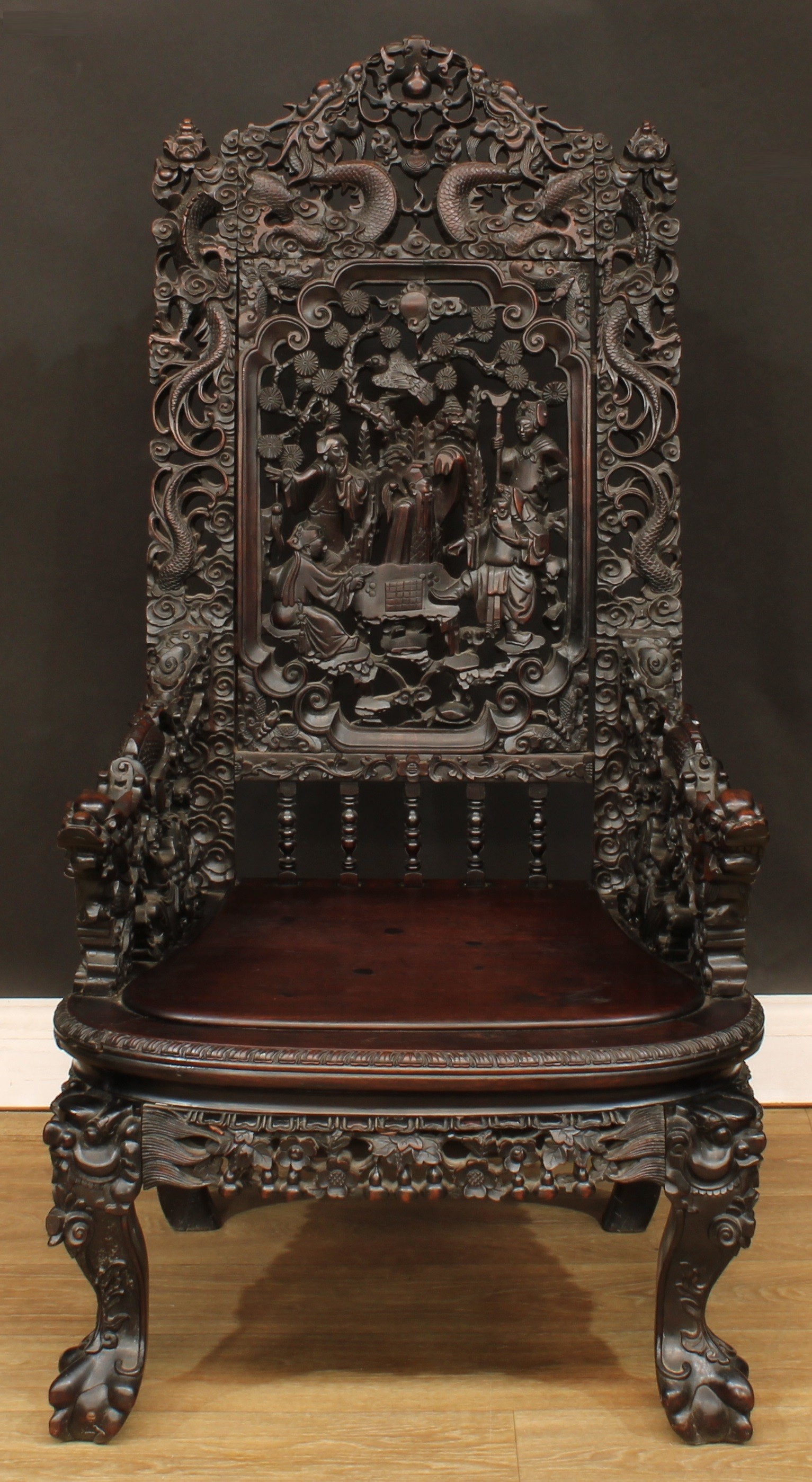 A Chinese hardwood armchair, shaped and pierced cresting carved with dragons, rectangular back panel carved with scholars partaking in a game of xiangqi beneath blossoming prunus, downswept arms carved as dragons, panel seat, cabriole forelegs, ball and claw forefeet, 114cm high, 60cm wide, the seat 48cm wide and 46cm deep