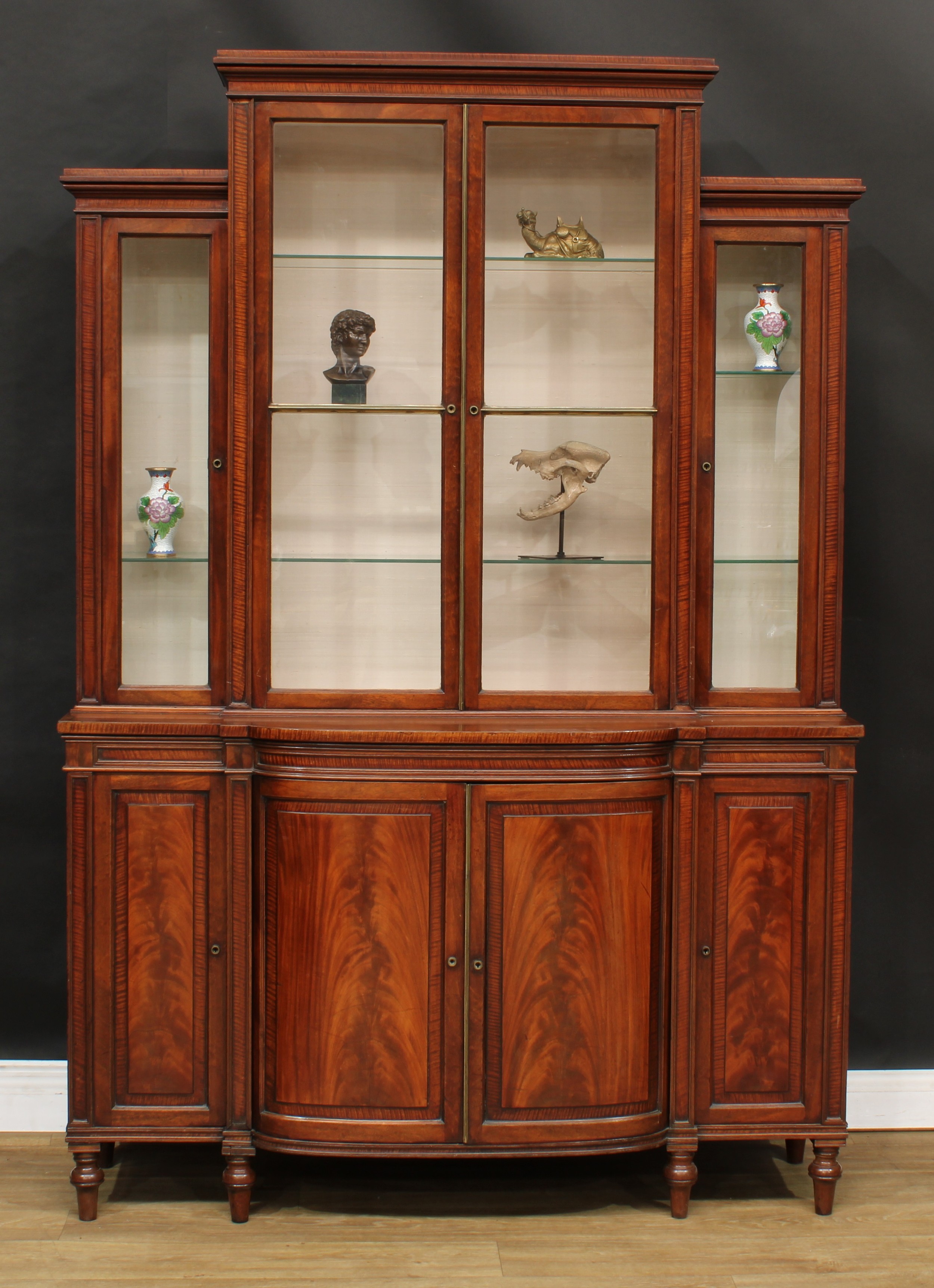 A 19th century fiddleback banded mahogany library bookcase, the breakfront superstructure with four glazed doors, the projecting bow-centre base with four panel doors, Bramah locks, turned feet, 196.5cm high, 137cm wide, 43cm deep