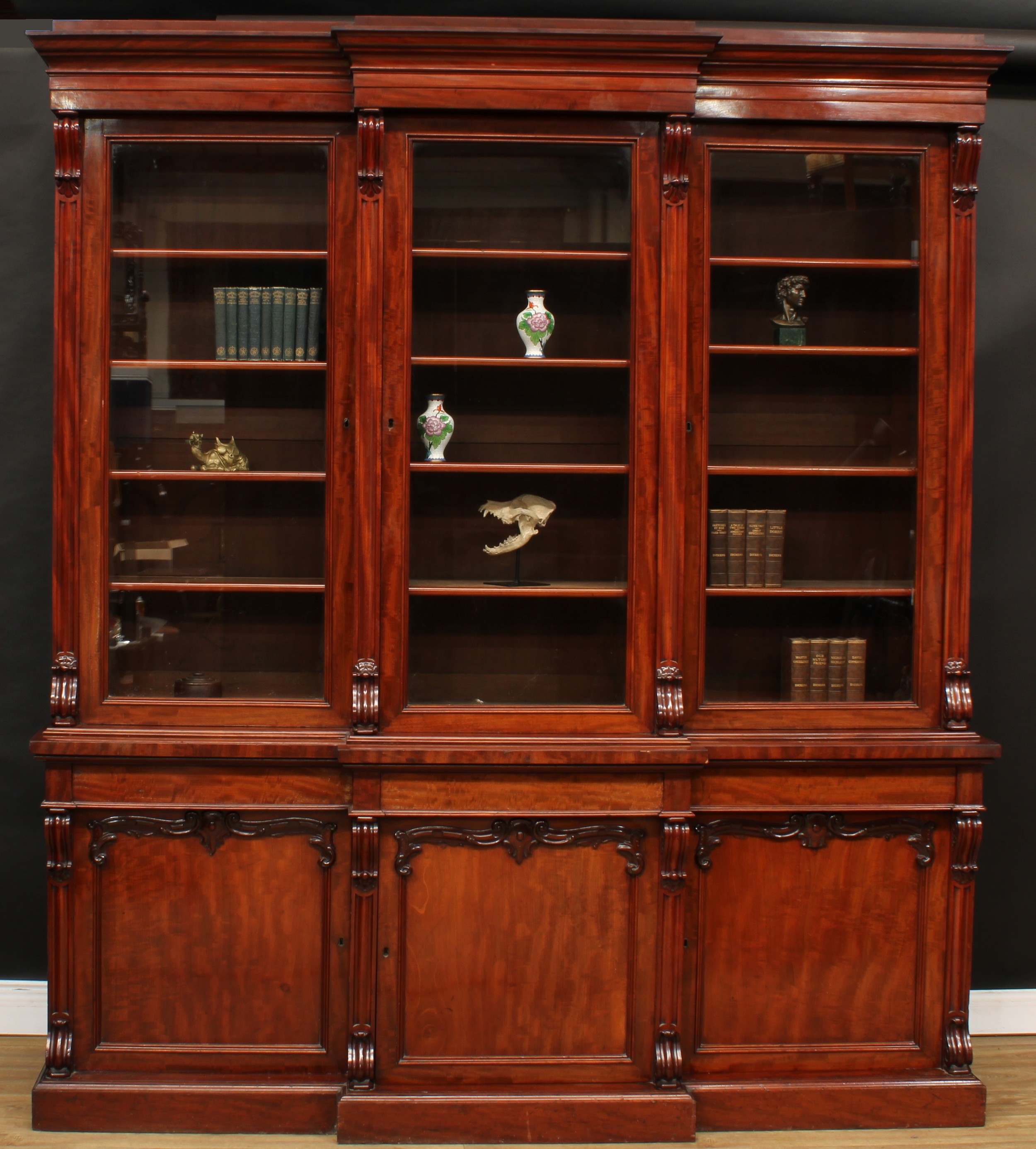 An early Victorian mahogany breakfront library bookcase, moulded cornice above three glazed doors, each enclosing four shelves, divided and flanked by scroll brackets, the projecting base with three panel doors, the left locked, the central enclosing a shelf, the right enclosing four drawers, plinth base, 243cm high, 217.5cm wide, 53.5cm deep, c.1845