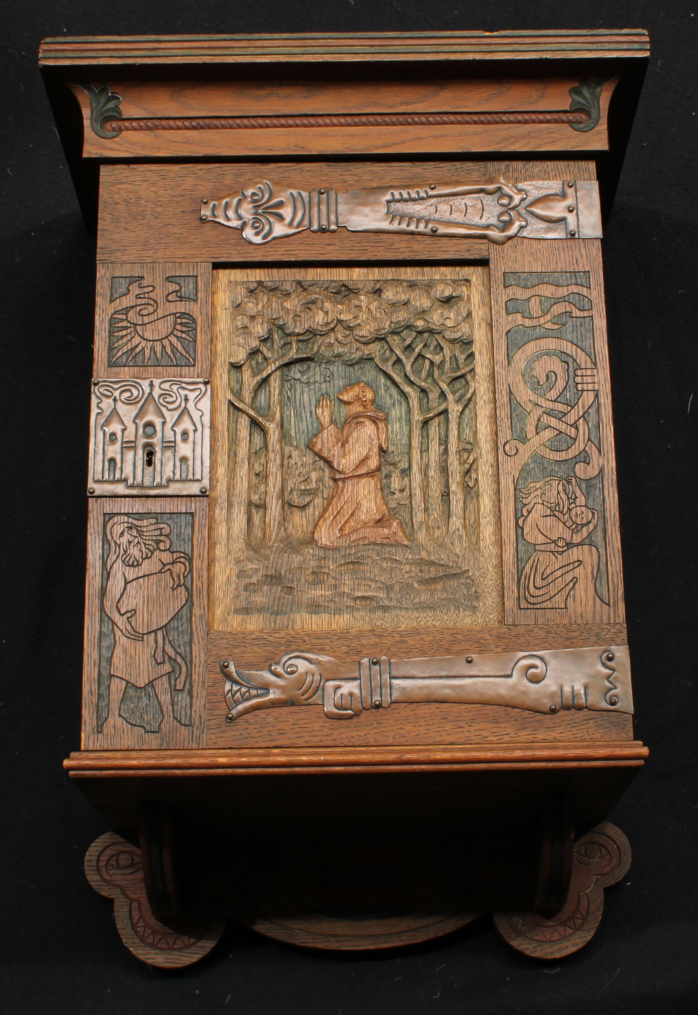 An Arts & Crafts oak wall hanging cabinet, outswept cornice above a rectangular door carved with figures, the central panel carved with a monk kneeling in prayer, zoomorphic strap hinges, the escutcheon embossed as a place of worship, the side indecipherably signed and dated 1906, shaped apron, 71cm high, 49cm wide, 28.5cm deep