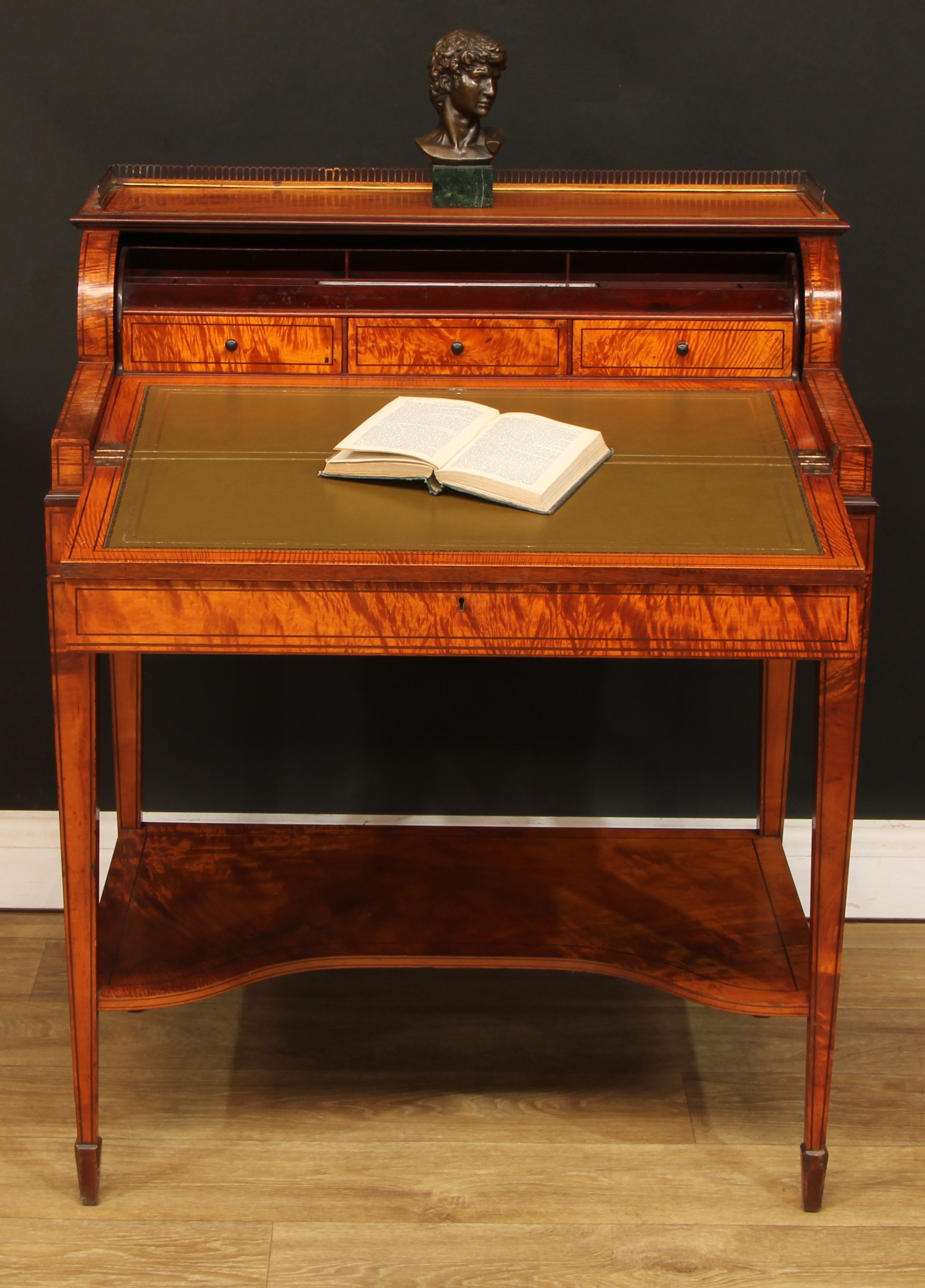 A Sheraton Revival satinwood and marquetry cylinder desk, by Hamptons, Pall Mall, London, rectangular top with brass three-quarter gallery above a retractable cylinder door, opening in conjunction with the frieze drawer, enclosing shallow pigeonholes, a pen tray and three small drawers, above a fold-out writing surface, the frieze drawer with Hobbs lock, tapered square legs, open undertier, spade feet, 93cm high, 75.5cm wide, 50cm deep, c.1900