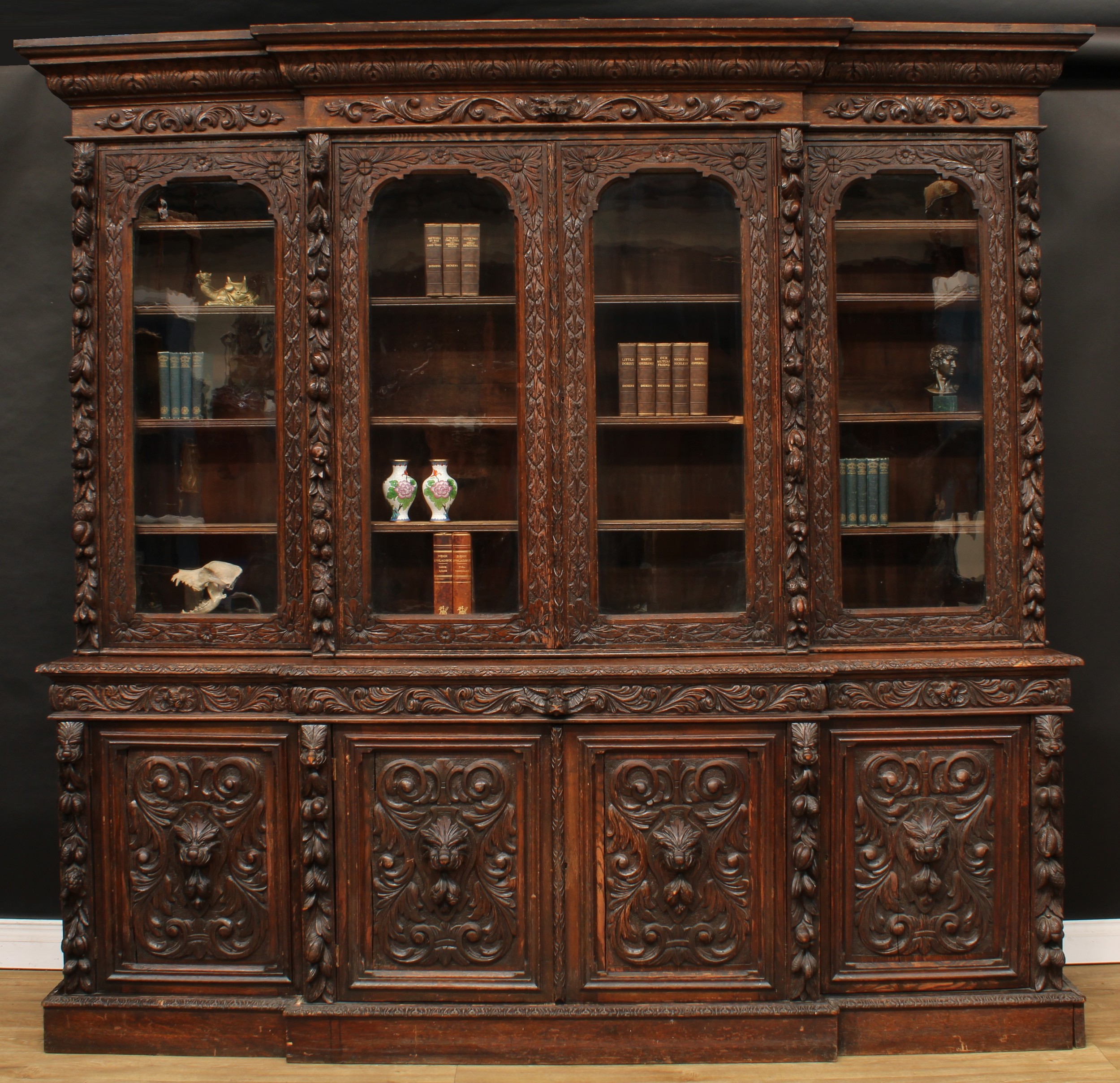 A substantial Victorian oak library bookcase, outswept cornice above four glazed doors enclosing shelves, the projecting base with four panel doors, carved throughout with fruit, flowers, leaves and leafy lion masks, plinth base, 241cm high, 259cm wide, 53cm deep, c.1880
