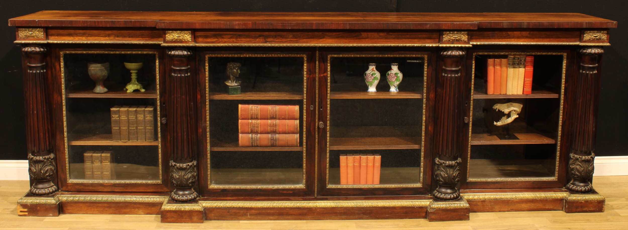 A large Post-Regency gilt metal mounted rosewood break-centre low library bookcase, possibly Gillows of Lancaster and London, slightly oversailing top above four glazed doors, divided and flanked by acanthus-grasped reeded pilasters, plinth base cast and applied with a gadrooned mount, 101.5cm high, 307cm wide, 55cm deep, c.1825