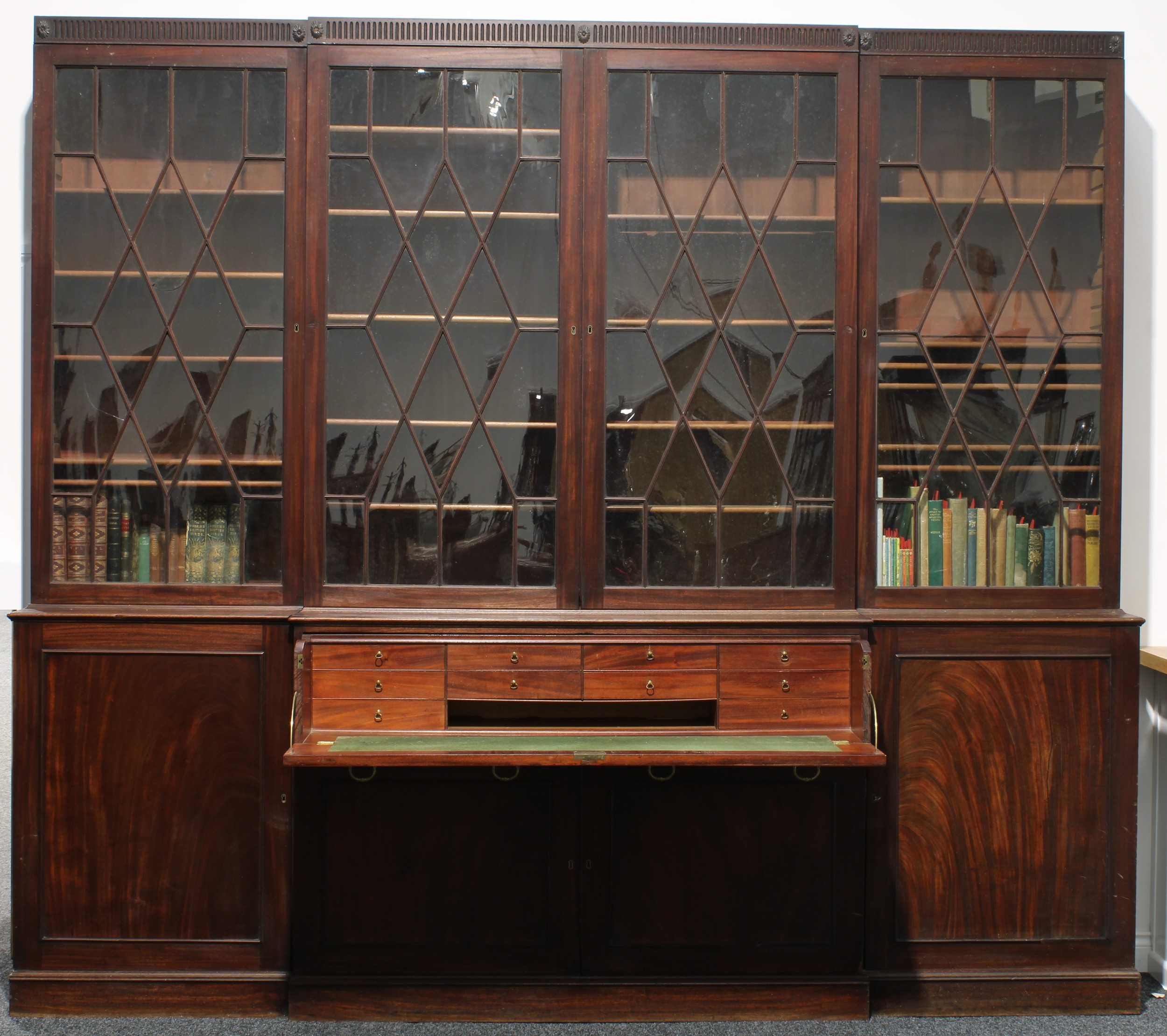 A substantial 19th century mahogany break-centre secretaire library bookcase, possibly Gillows of Lancaster and London, nulled capital carved and applied with flower head bosses above four astragal glazed doors, each enclosing adjustable shelves, the projecting base with a long drawer with Bramah lock, falling for writing and enclosing small drawers, above and flanked by panel doors, 265cm high, 301cm wide, 53.5cm deep