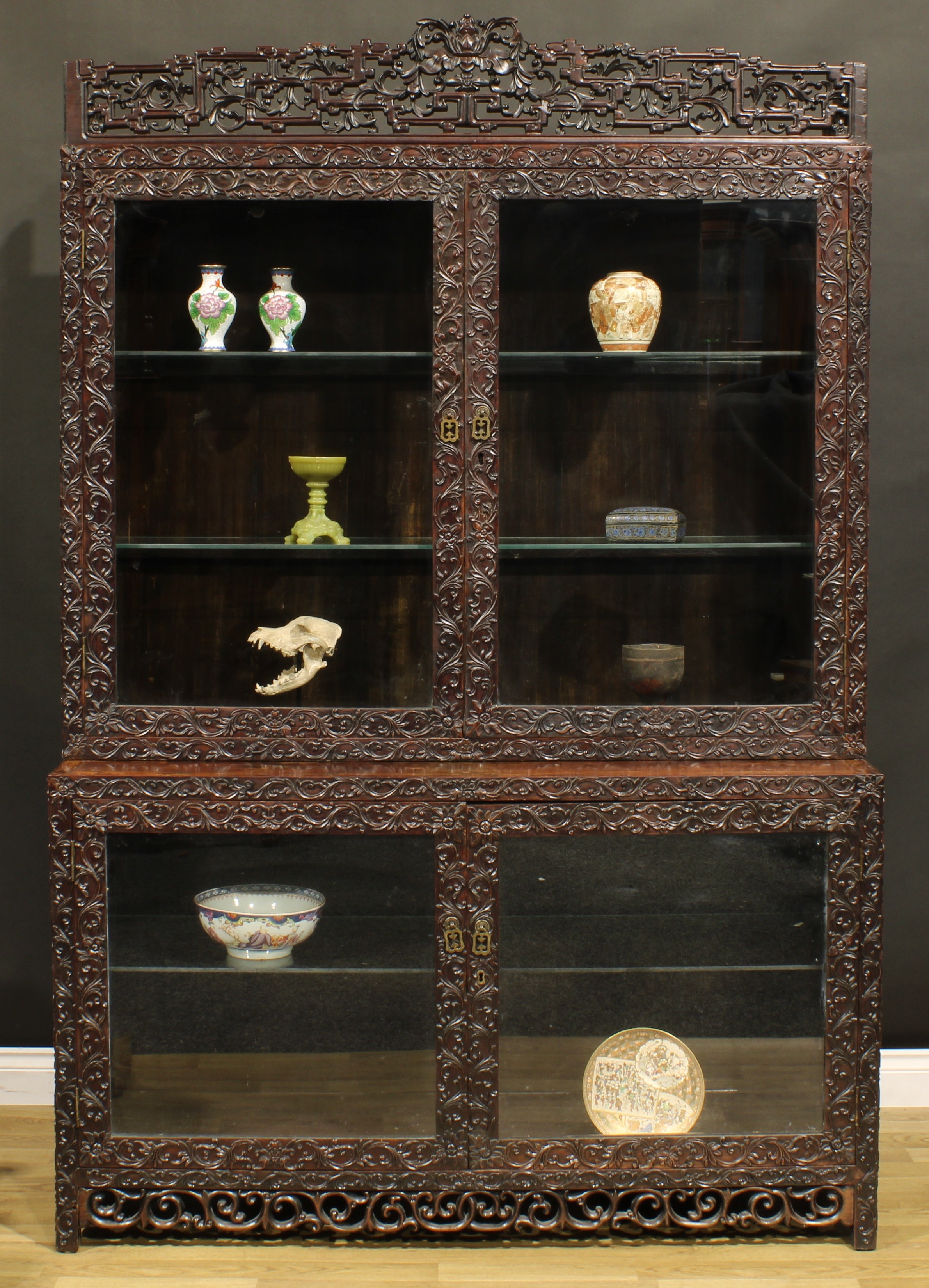A Chinese hongmu porcelain vitrine or display cabinet, shaped pediment pierced and carved with meandrous leaves and stylised angular clouds, above a pair of glazed doors enclosing two shelves, the projecting base with a further pair of doors enclosing a shelf, carved throughout with flower heads and meandrous foliage, 223cm high, 140.5cm wide, 51cm deep