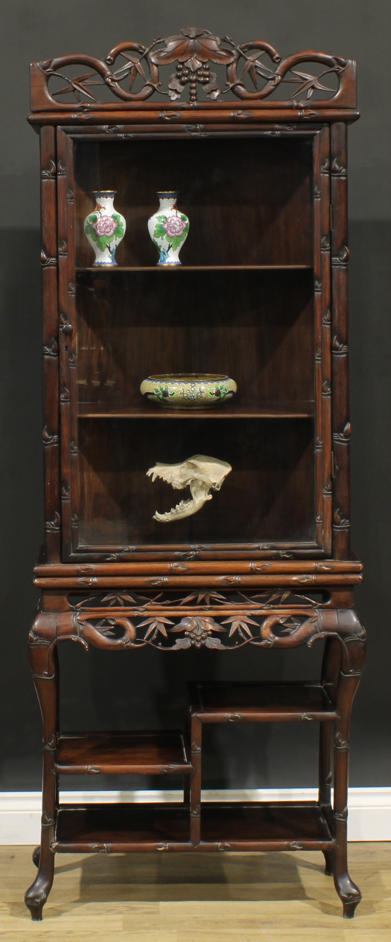 A Chinese hardwood porcelain vitrine or display cabinet, shaped pediment pierced and carved with fruiting vine above a glazed door enclosing two shelves, the base with an asymmetrical arrangement of shelves, carved throughout with leafy bamboo, 180cm high, 67.5cm wide, 31.5cm deep