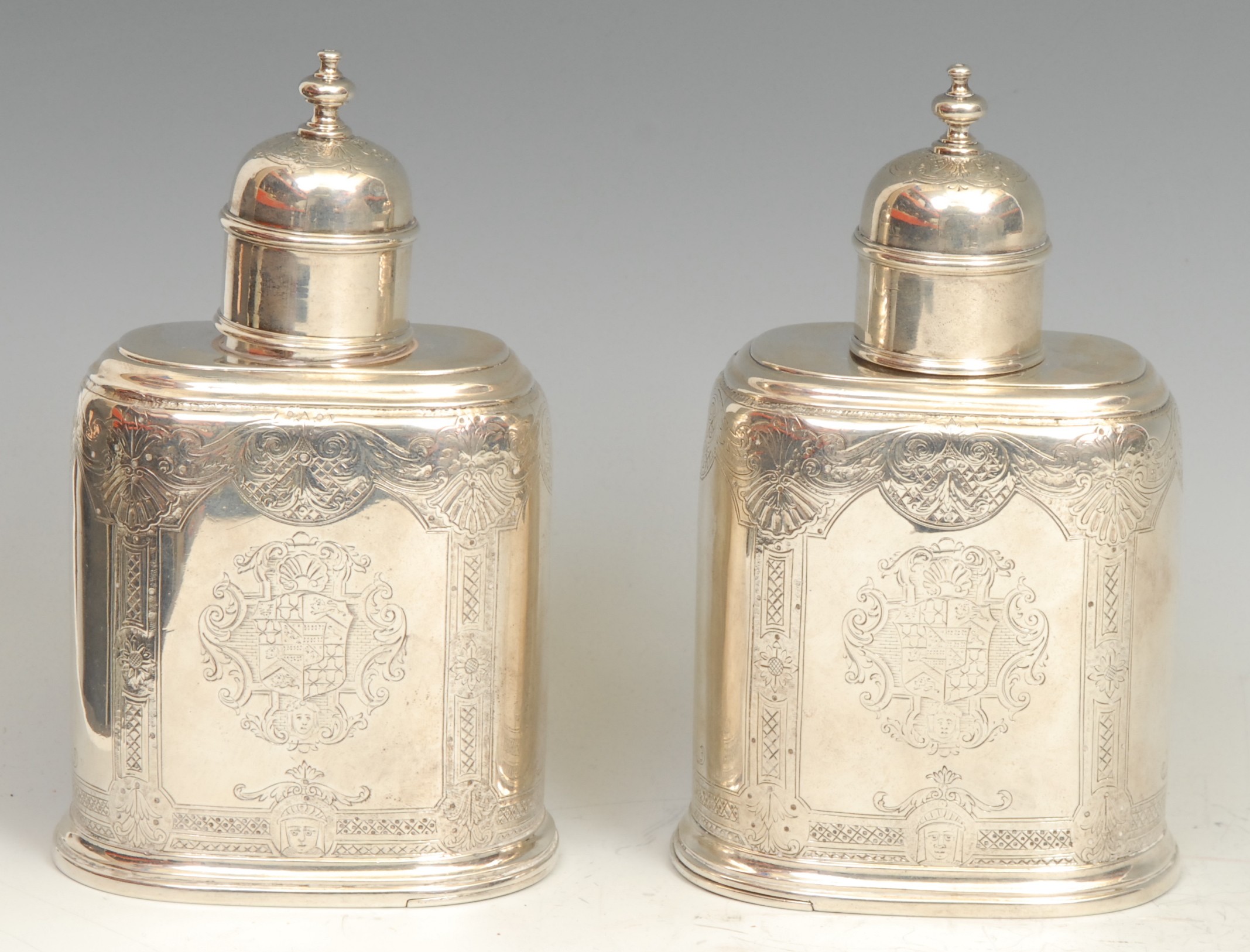 A pair of early George II silver oval tea caddies, bell-shaped push-fitting covers, flat-chased with Rococo borders and engraved with masks, sliding covers to bases, 14cm high, struck with lion passant and maker three times, John Newton, London c.1730, 494g
