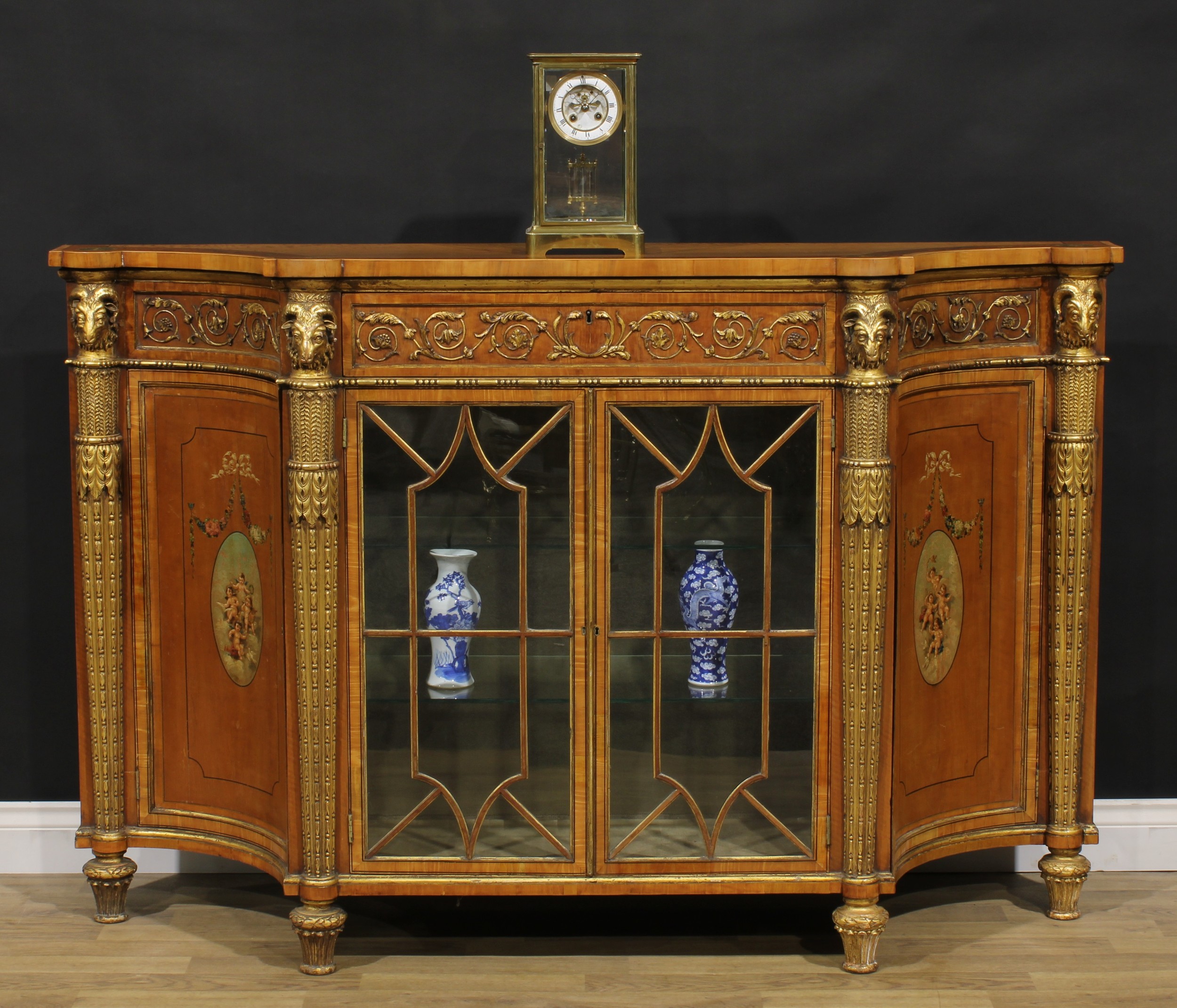 An Adam Revival parcel-gilt satinwood and painted side cabinet, slightly oversailing top above a single frieze drawer and a pair of astragal glazed doors enclosing two shelves, flanked by concave panel doors, each enclosing a further shelf, divided and flanked by ram-capped engaged columns, applied with leaves and bell husks, lotus feet, 107cm high, 165cm wide, 43cm deep