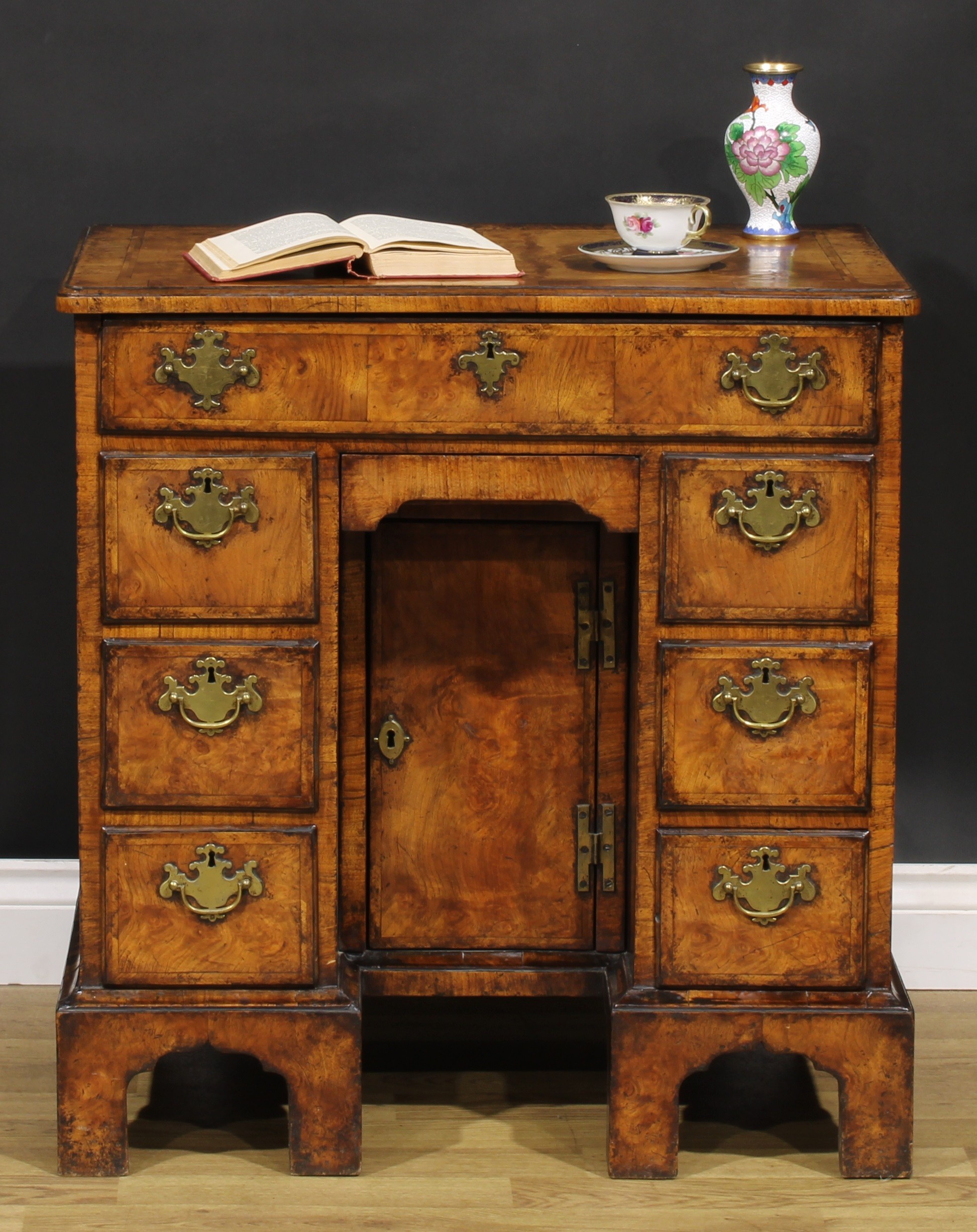 A George I/II walnut kneehole desk, of small proportions, quarter-veneered and crossbanded top with moulded edge above a long drawer, an apron drawer, and a cupboard, flanked by further drawers, outlined throughout with festherbanding, brass swing handles, batwing backplates, tall bracket feet, 77cm high, 72cm wide, 44cm deep, c.1730