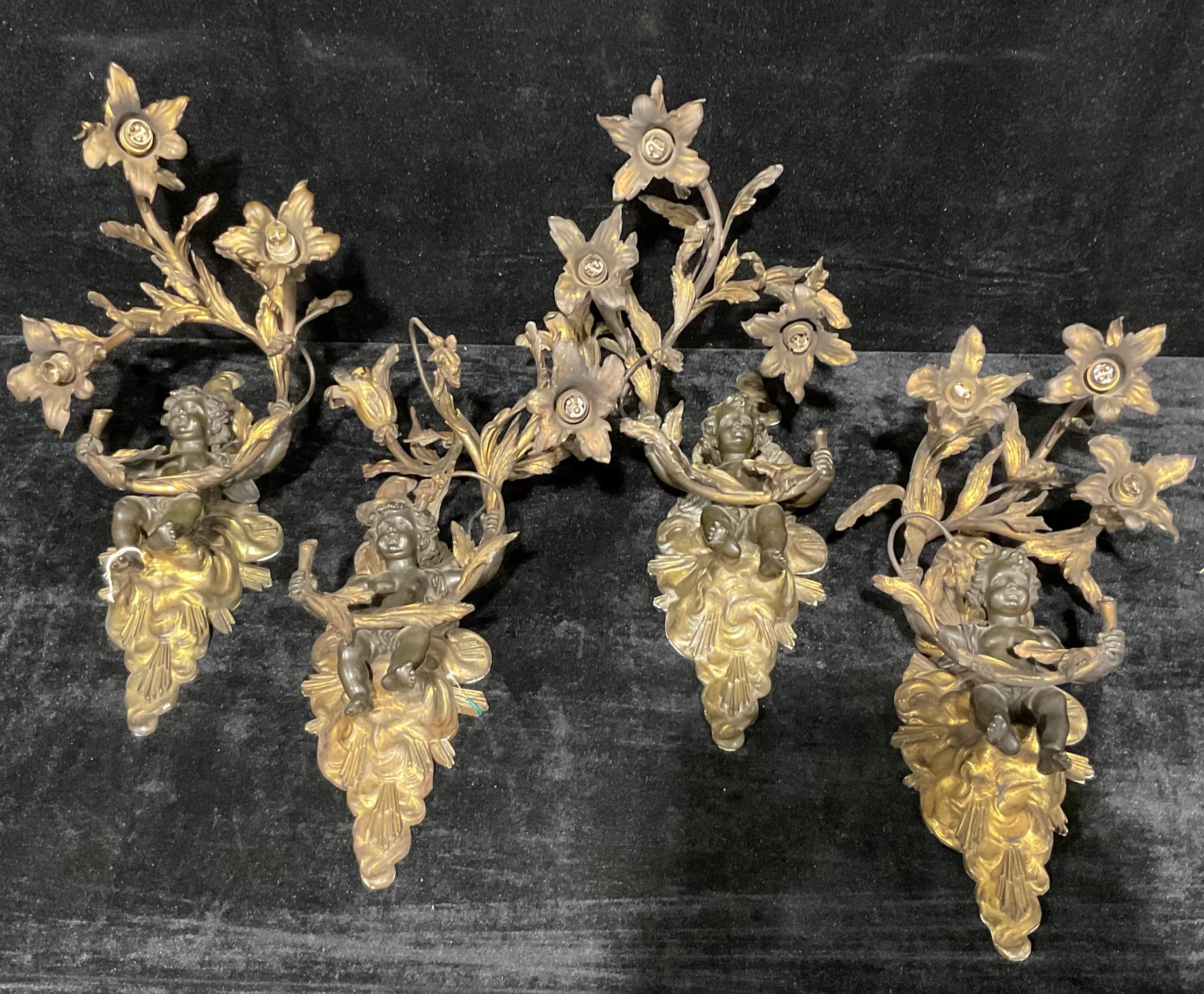 A suite of four Louis XV Revival dark patinated and parcel-gilt bronze three-light electrolier wall sconces, each cast as a putto amongst flowering branches, the mounts with sun rays bursting through clouds, 45cm high, early 20th century