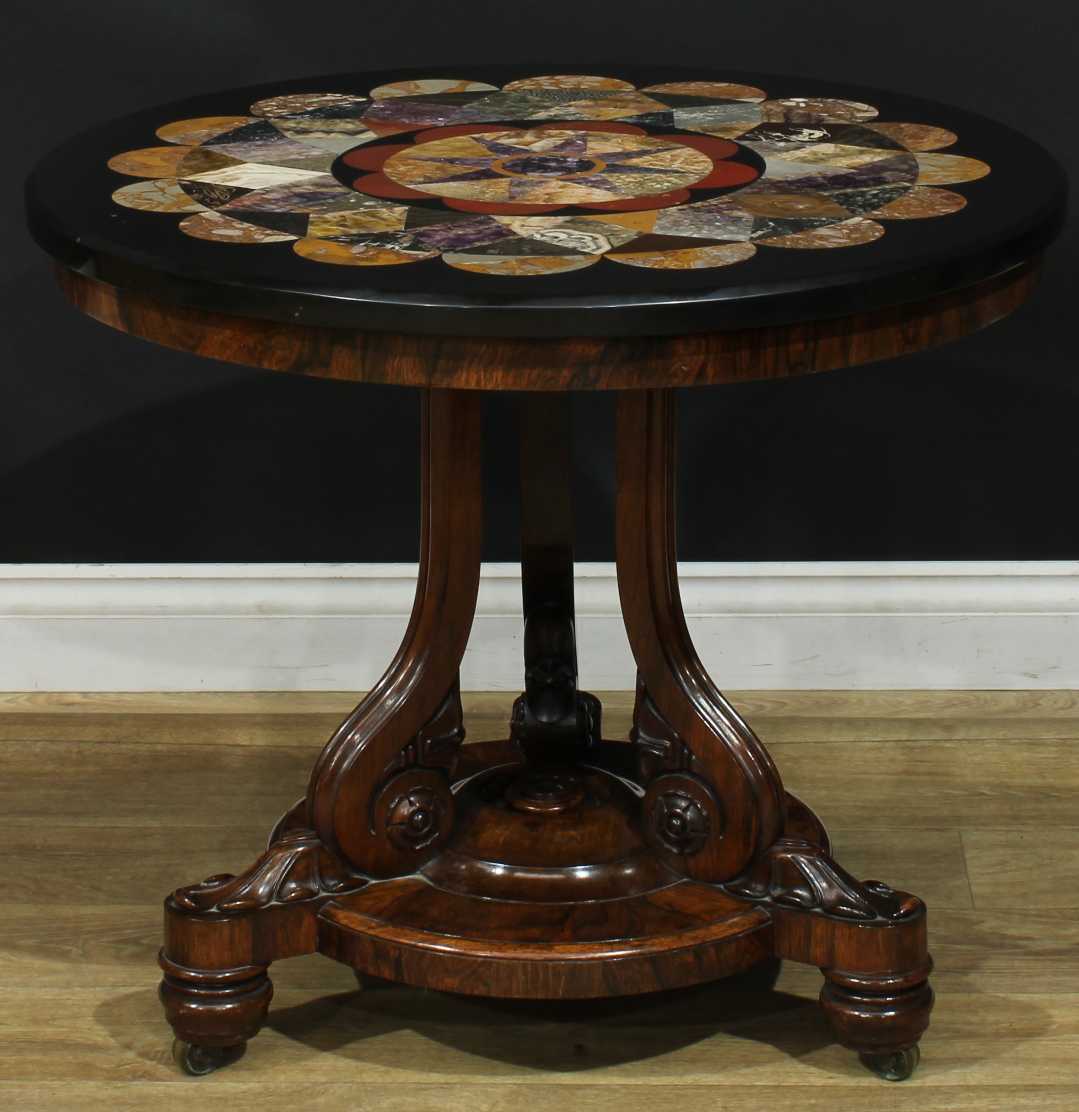 A Post-Regency Derbyshire Ashford marble mounted rosewood centre table, circular top inlaid in Blue John, fossil stone, Carnelian and other specimens samples, with bands of lozenges and demilunes centred by an eight-pointed star, the base in the typical William IV taste with three scroll supports, turned feet, brass casters, 66cm high, the top 76cm diameter, the elements mid-19th century