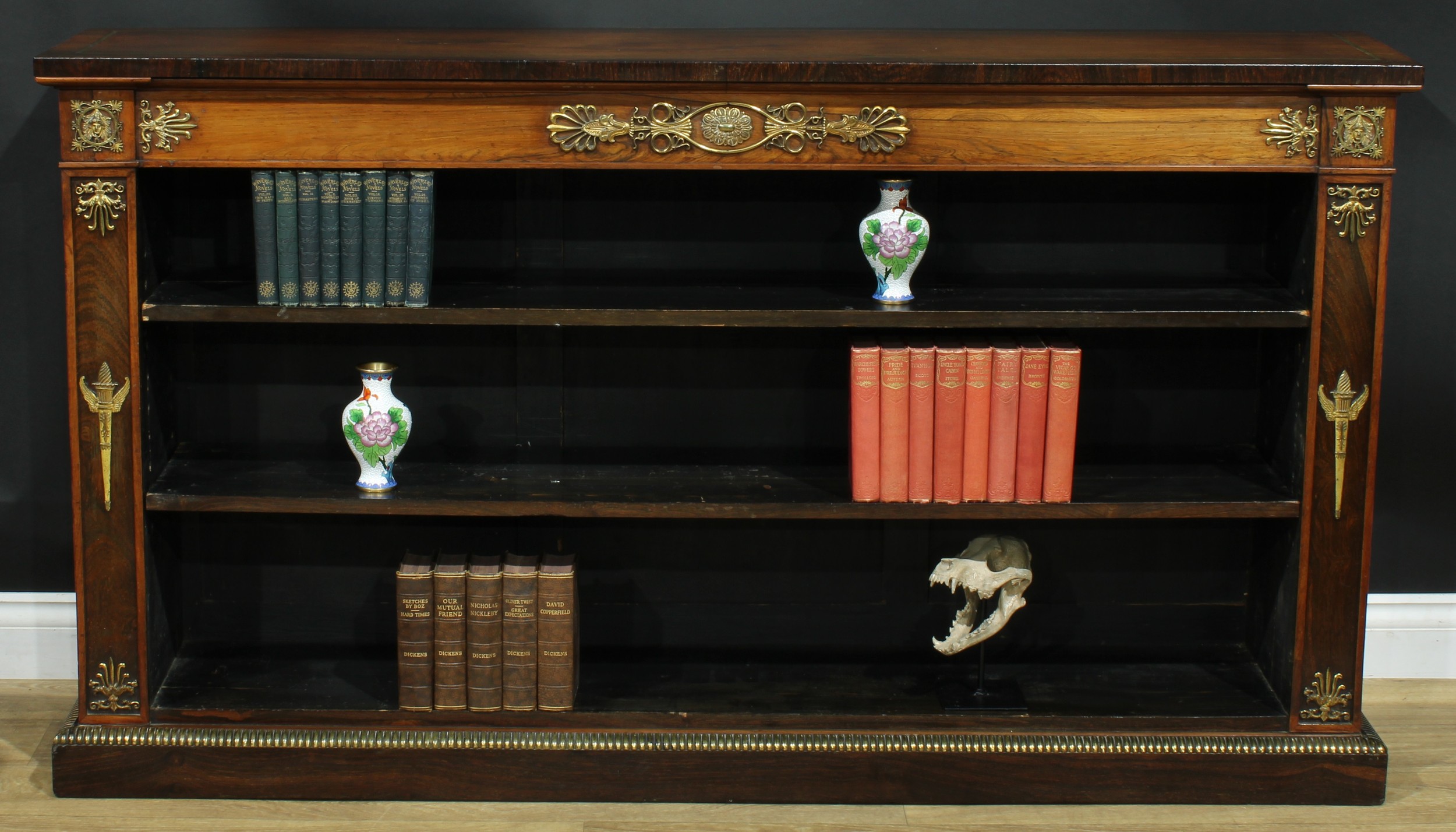 A Regency gilt metal mounted rosewood open library bookcase, oversailing top outlined with brass stringing above a deep frieze and two adjustable shelves, the borders applied with Classical devices, plinth base, 91.5cm high, 163.5cm wide, 34cm deep, c.1815
