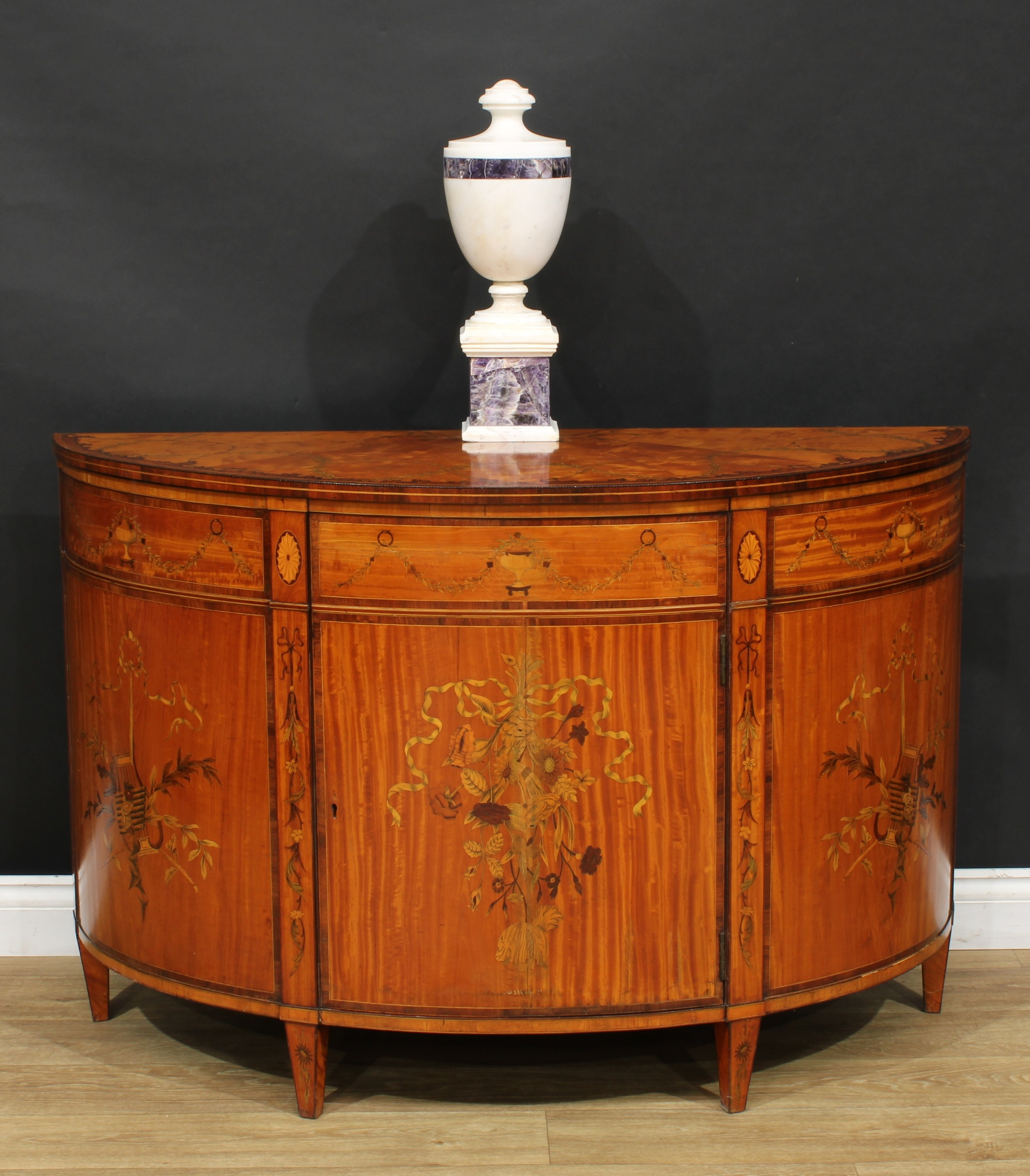 A Sheraton design satinwood and marquetry demilune side cabinet, rosewood crossbanded top with radiating veneers inlaid with meandrous leafy stems, a rear-central batwing patera and bell husks, the front edge with a wavy border of amboyna and outlined with barberpole stringing, above a rectangular door inlaid with a ribbon-tied spray of flowers, flanked by panels, each inlaid with a musical trophy, divided and flanked by further ribbon-tied stems, tapered square legs, 81cm high, 122cm wide, 48cm deep, 19th century