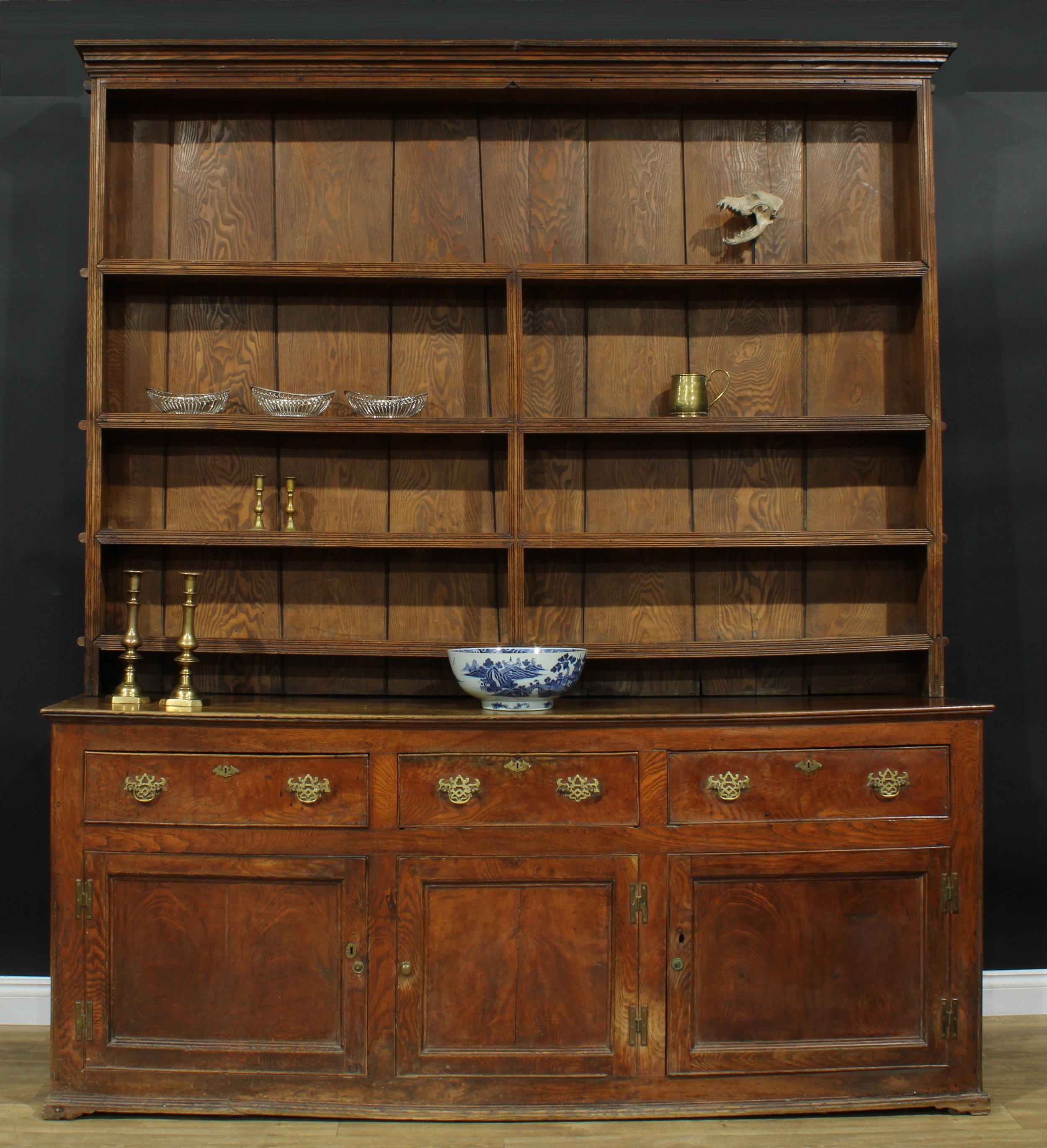 A large George III ash and elm country house dresser, outswept cornice above four plate rack shelves, the projecting base with slightly oversailing top above three frieze drawers and three panel doors, 250cm high, 218cm wide, the base 91cm high, 213.5cm wide and 56cm deep, c.1780