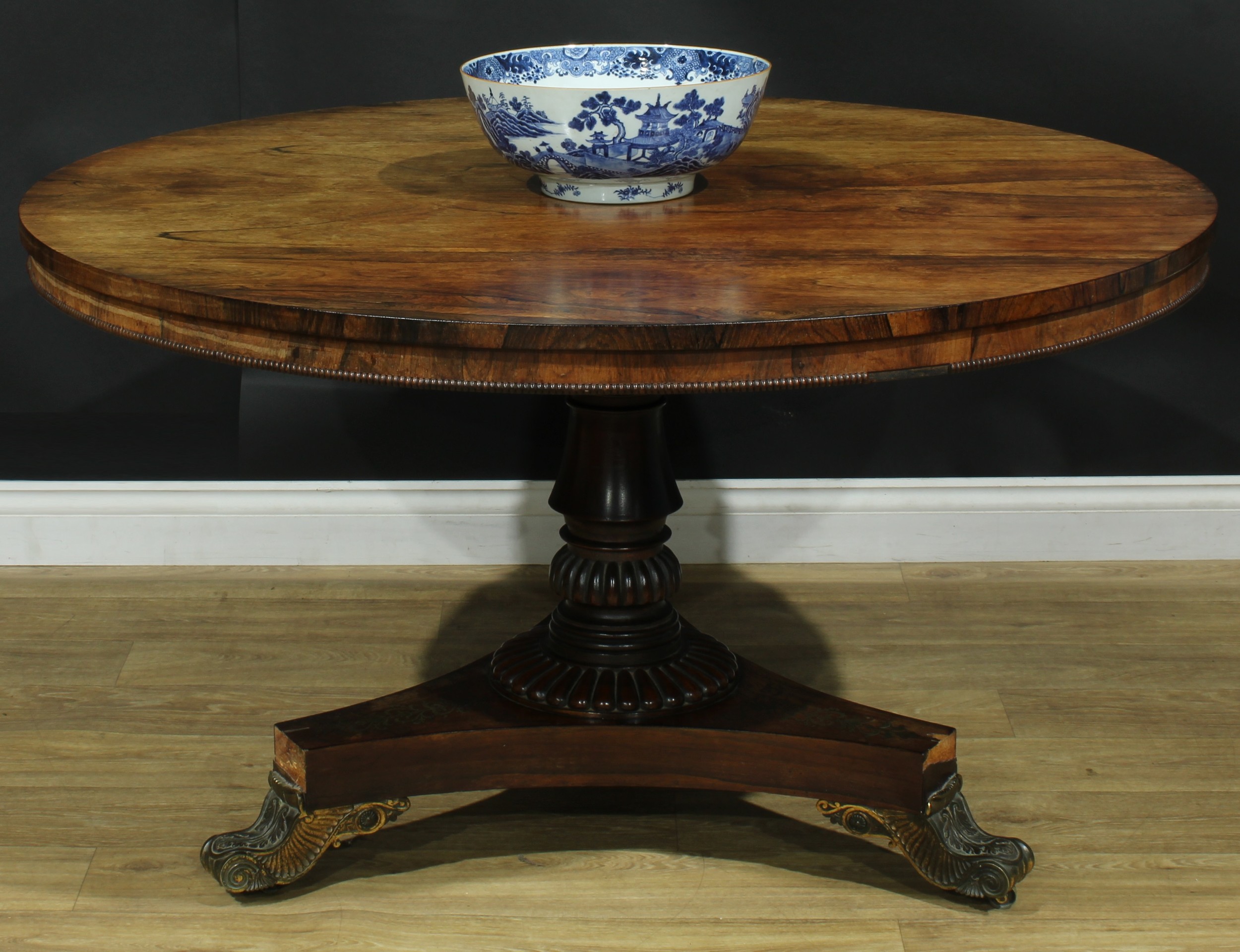 A Post-Regency gilt metal mounted rosewood and brass marquetry centre table, circular tilting top, turned column, gadrooned socle, incurve triform base inlaid with stylised leaves and bell husks, scroll feet cast with acanthus, brass casters, 72cm high, 125.5cm diameter, c.1830