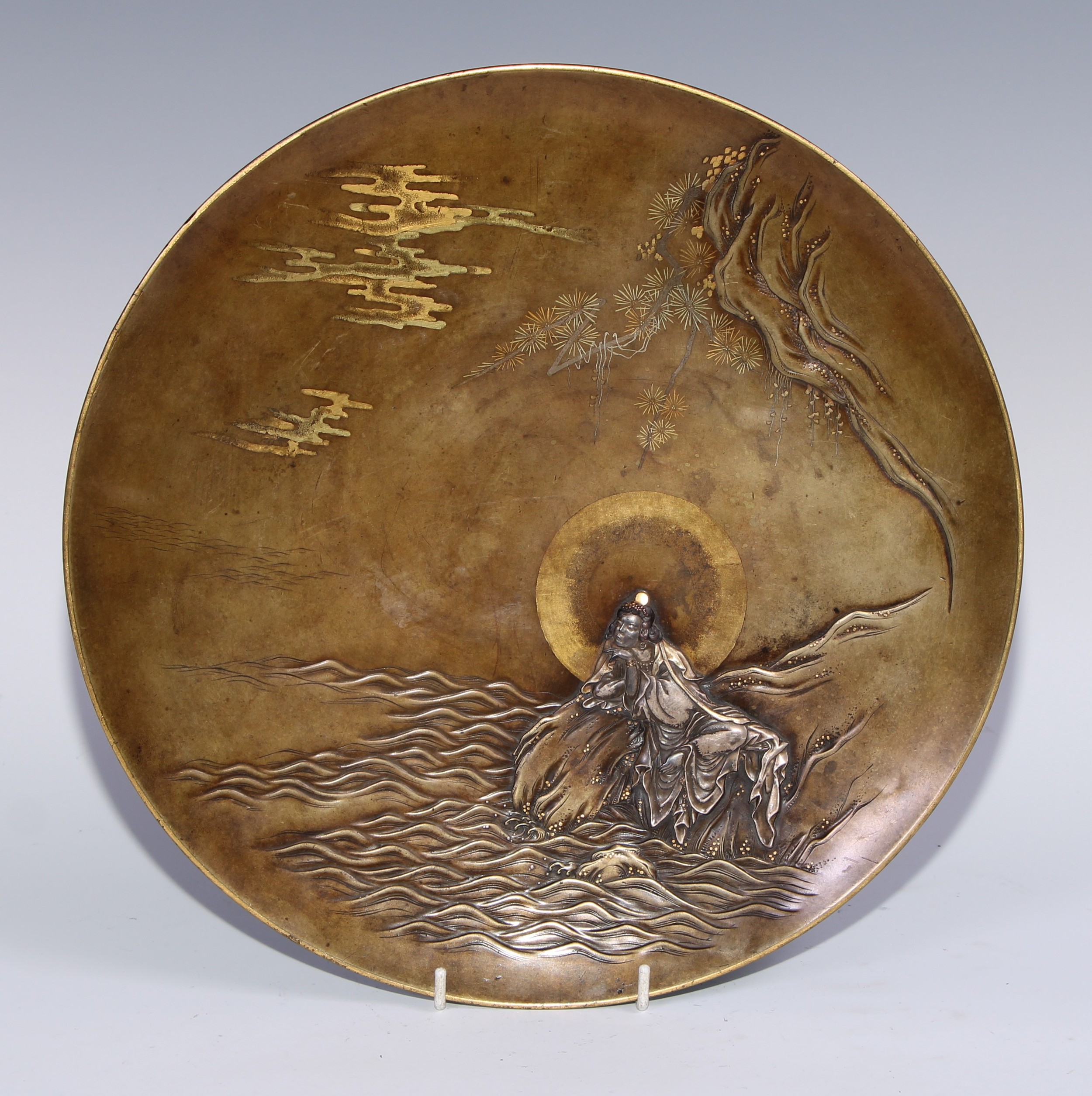 A Japanese bronze circular charger, applied and inlaid in silver and mixed metal with Guanyin on a rock at the edge of the coast, 32.5cm diam, signed and inscribed to verso, 32.5cm high, Takaoka, Meiji period