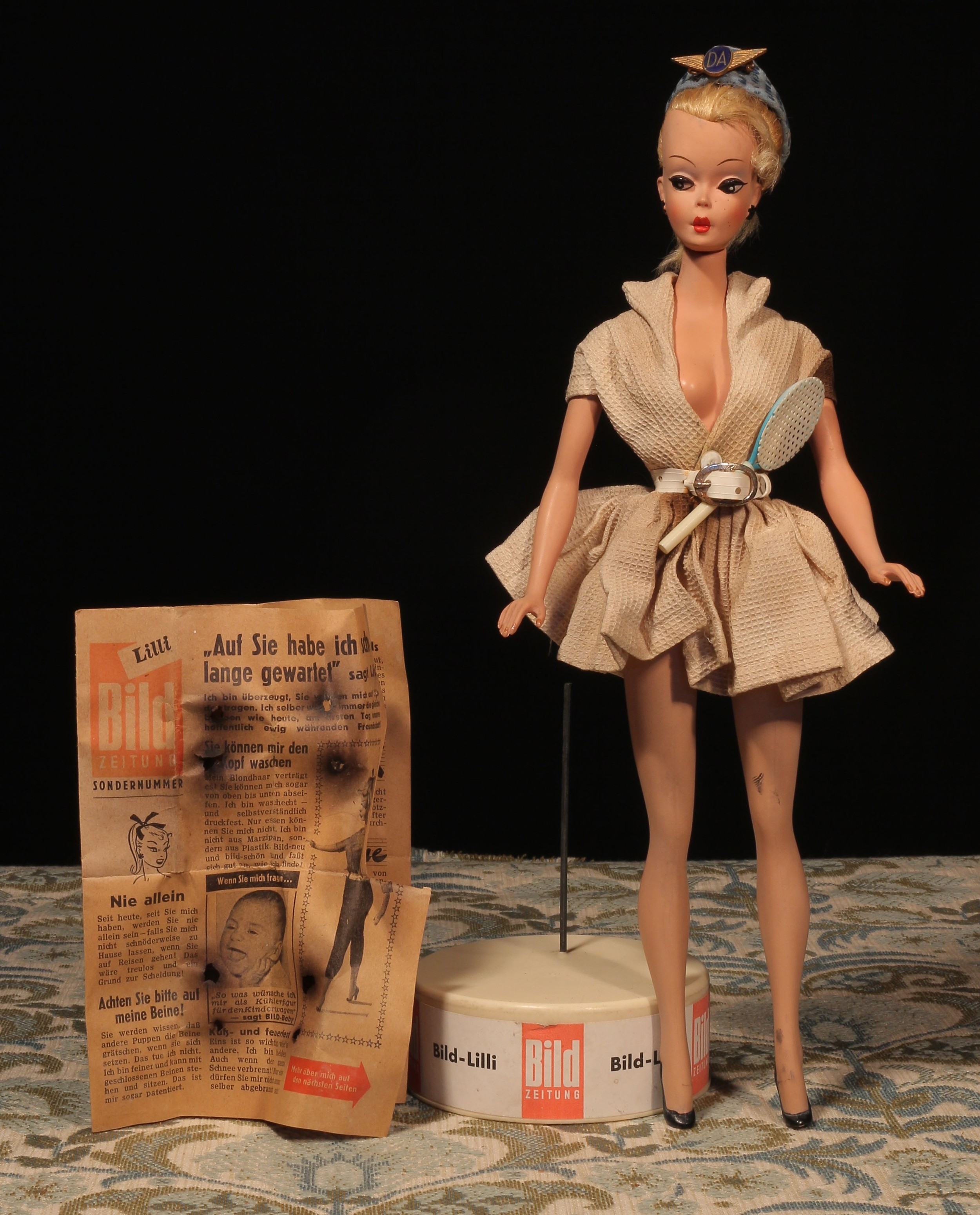 A rare 1950's or 1960's Bild-Lilli (Germany) 11½" jointed plastic doll, the swivel head with painted features including brown eyebrows, black side glancing eyes and red lips, blonde ponytail hair, black moulded and painted earrings, wearing a dress with 'PRYM' pop stud, moulded and painted black high heel shoes with holes to undersides, 29cm high, unmarked; together with raised circular plastic stand applied with 'BILD-LILLI, BILD ZEITUNG' red and white paper label and miniature faux literature newspaper