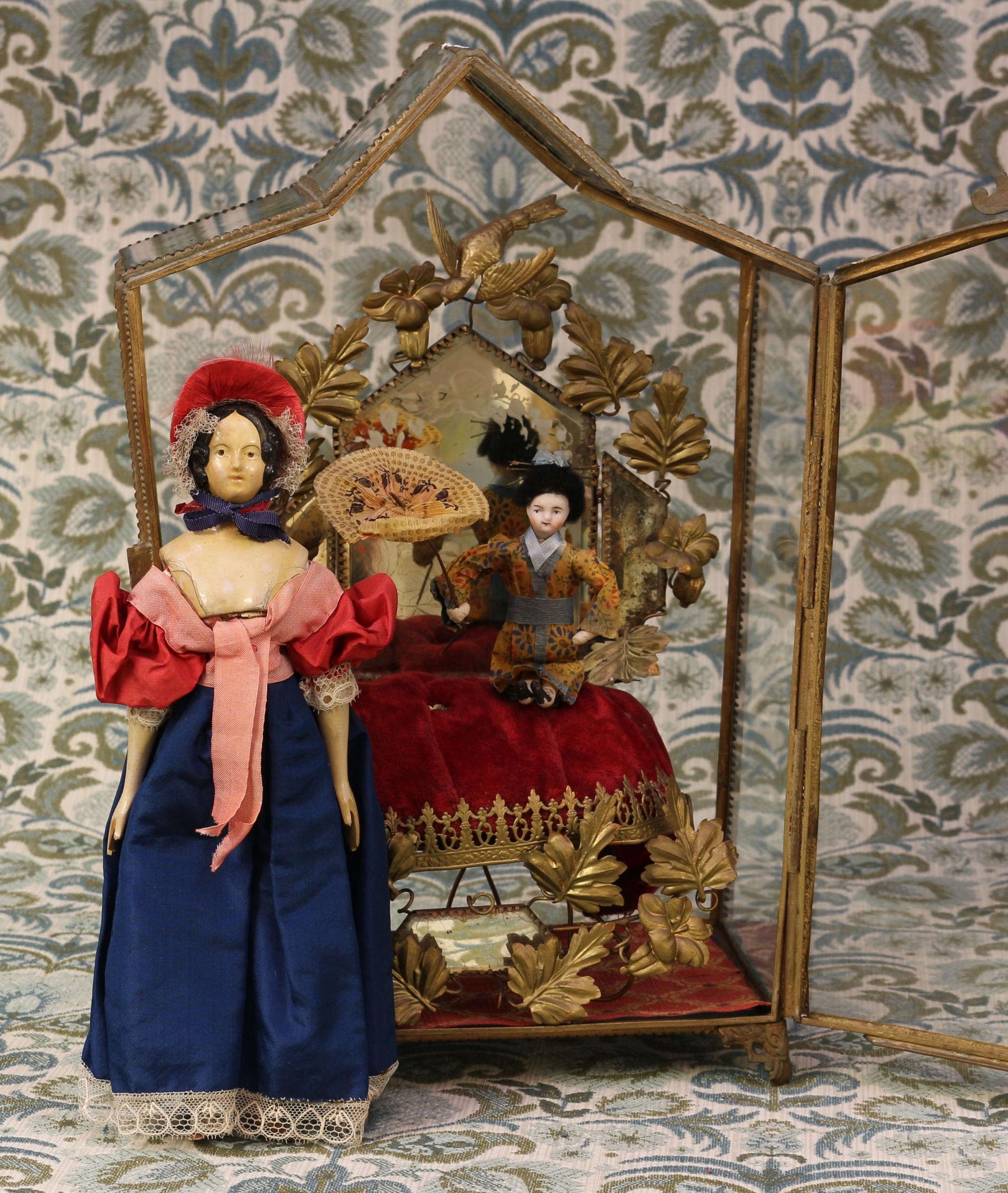 A mid 19th century painted papier-mâché shoulder head doll, the papier-mâché head with black moulded hair and painted features including brown eyes, partially kid leather body with narrow waist and wide flaring hips, carved and painted wooden arms and legs with shaped feet, 27cm high; a miniature pale bisque head doll, dressed as a Geisha, 13cm high; together with a French gilt brass glazed miniature display case or cabinet, with fitted interior, 39cm high, 27cm wide, 14cm deep (3)