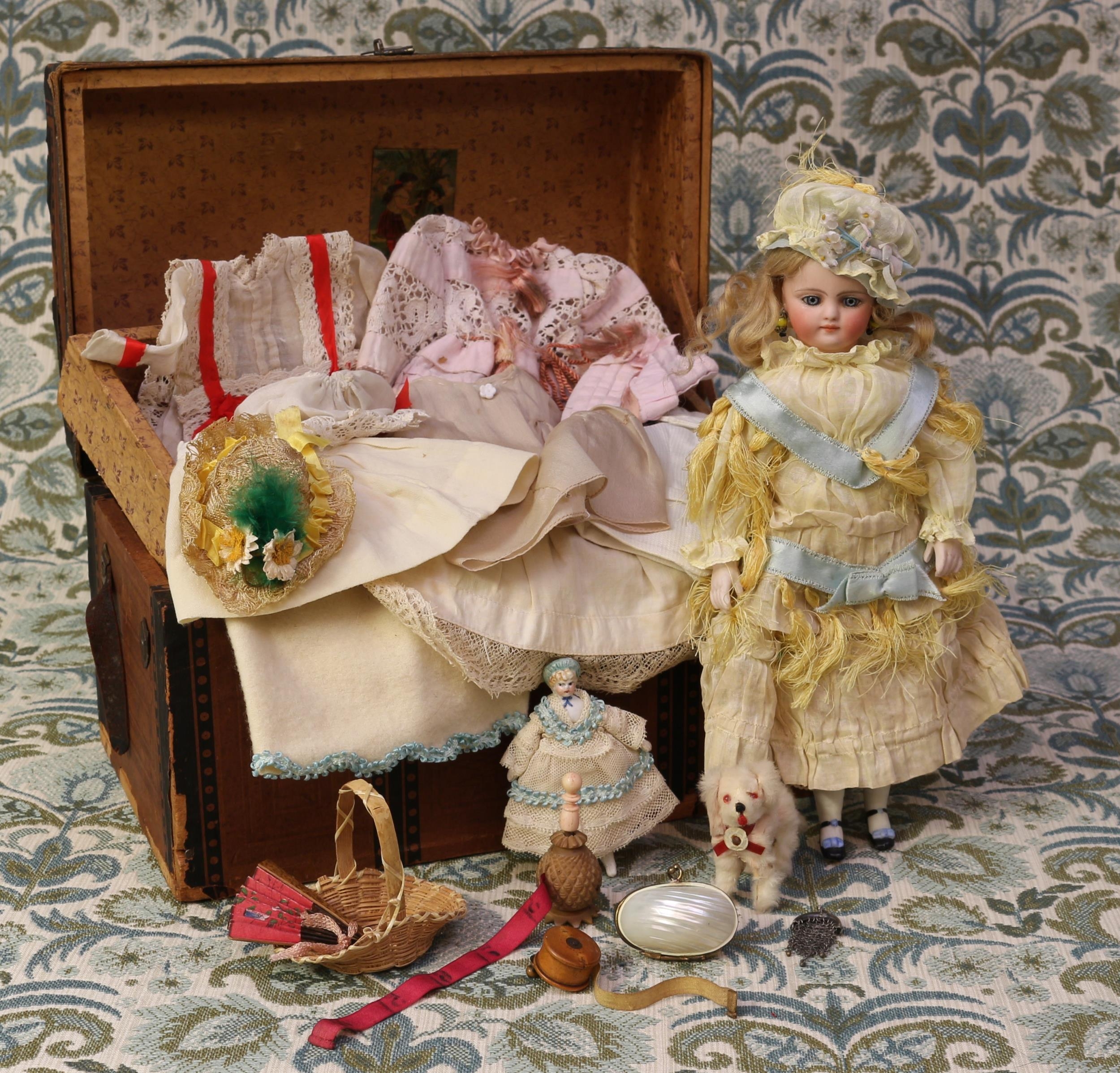 A late 19th century bisque shoulder and bisque head doll, of small proportions, the bisque head inset with fixed blue glass eyes and painted features including brown feathered eyebrows, fine eyelashes, red lips and blush cheeks, blonde wig on pate, pierced ears, partially stuffed cloth body with unglazed lower arms and legs, 27cm high; together with a rectangular shaped paper covered domed top trunk, of small proportions, 19.5cm high, 30.5cm wide, 18.5cm deep, the interior partially fitted with a lift out tray containing various doll clothing, accessories and miniatures (quantity)